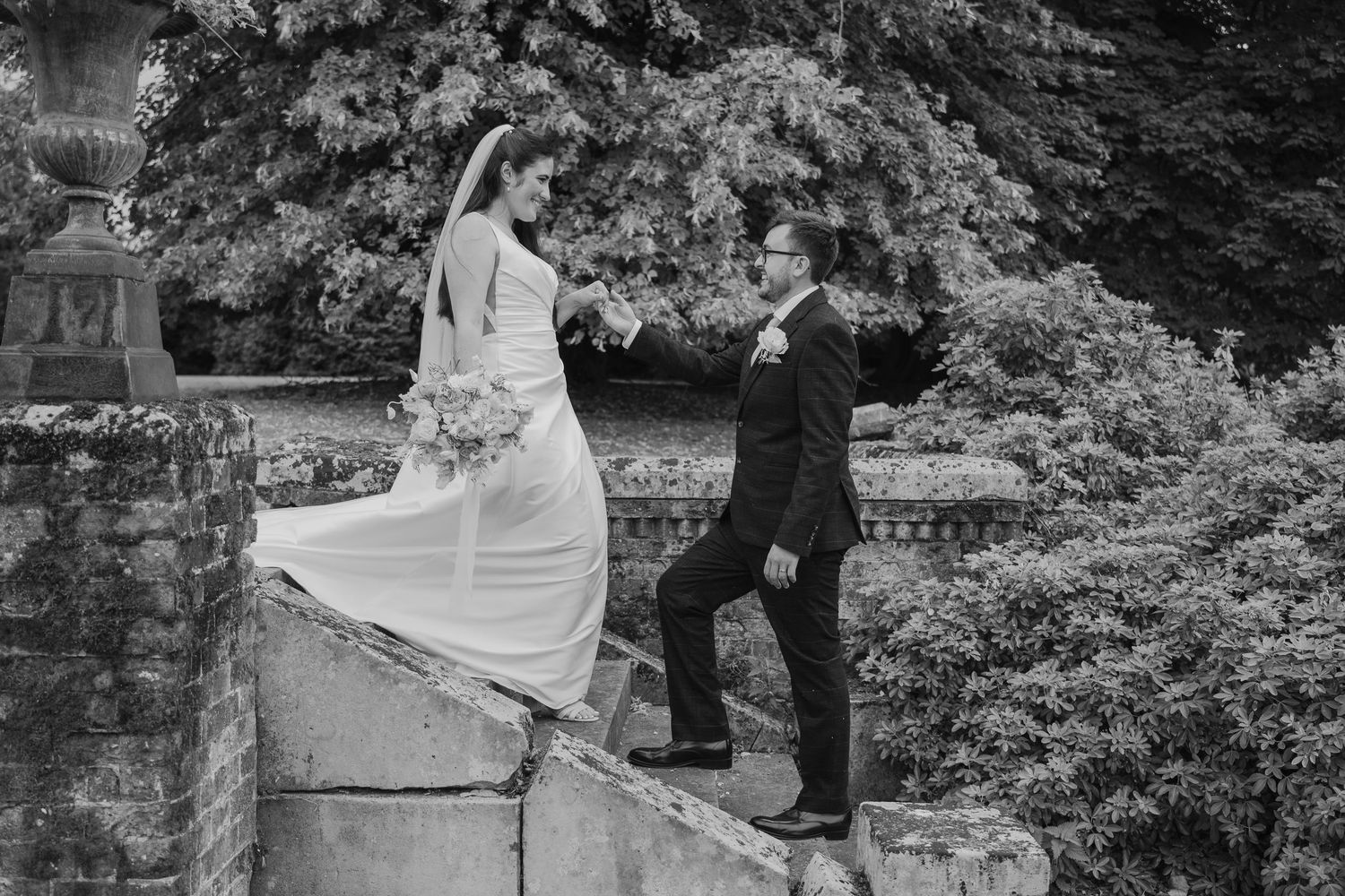 Black and white photo of bride and groom posing on stone steps in a lush garden, bride holding bouquet.
