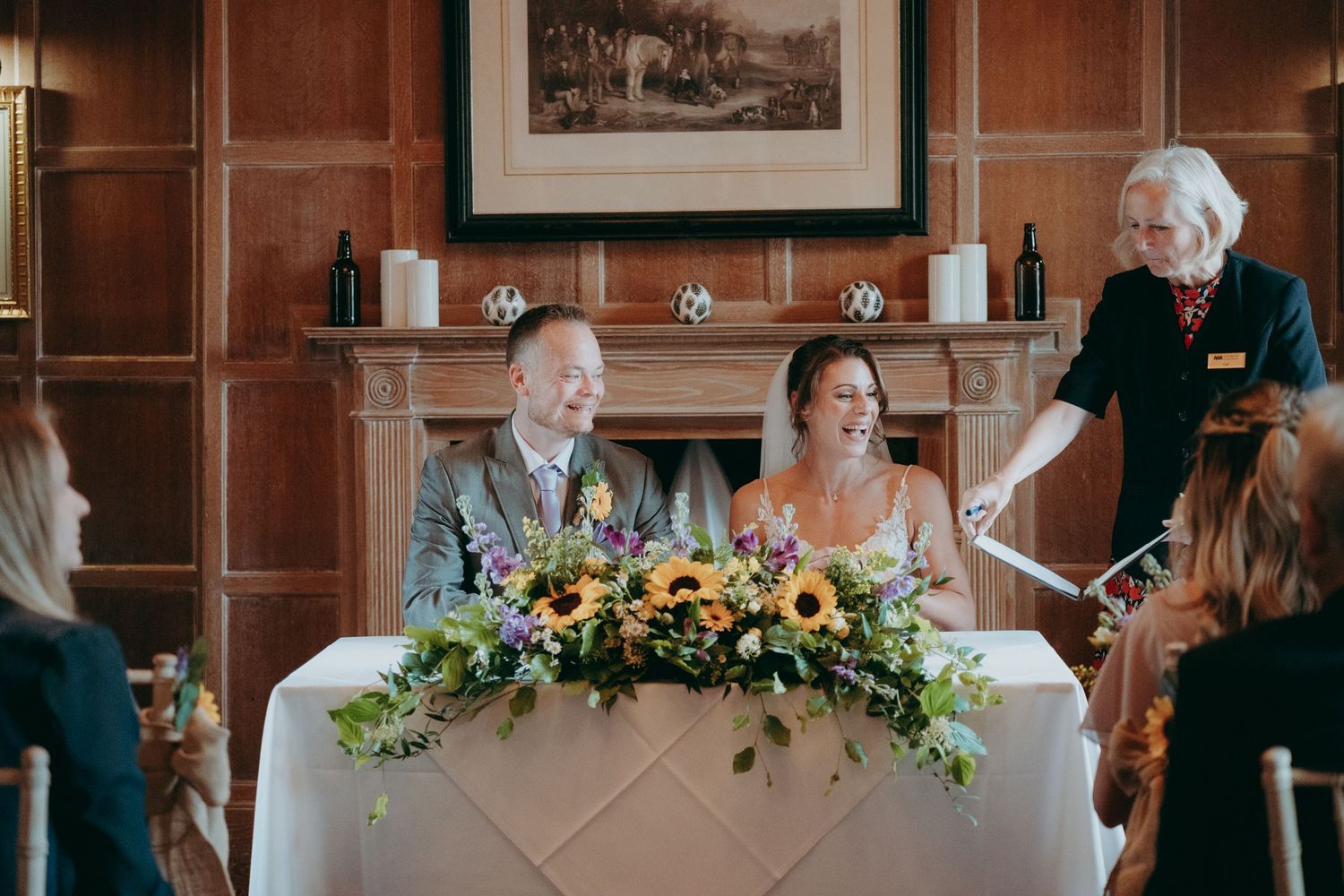 Couple smiling at wedding table with sunflower centerpiece, officiant beside them in elegant wood-paneled room.