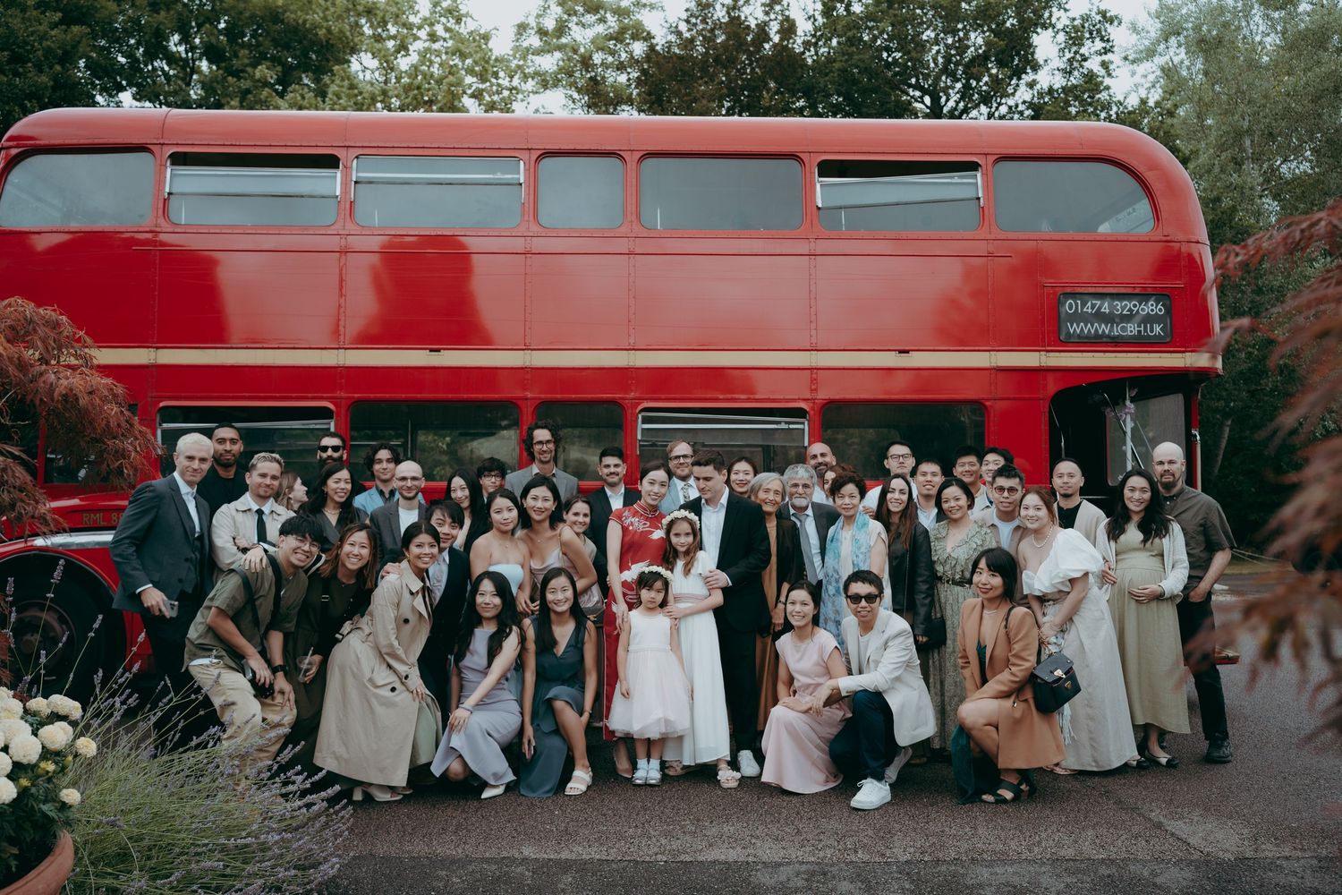 Large wedding group posing in front of a classic red double-decker bus outdoors.