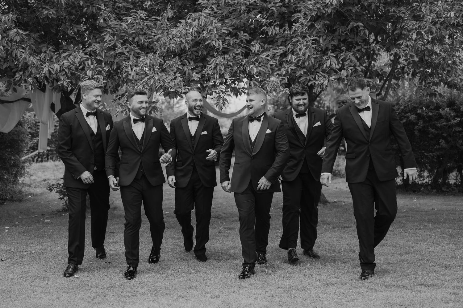 Black and white photo of a groom and groomsmen in tuxedos walking together outdoors at a wedding.