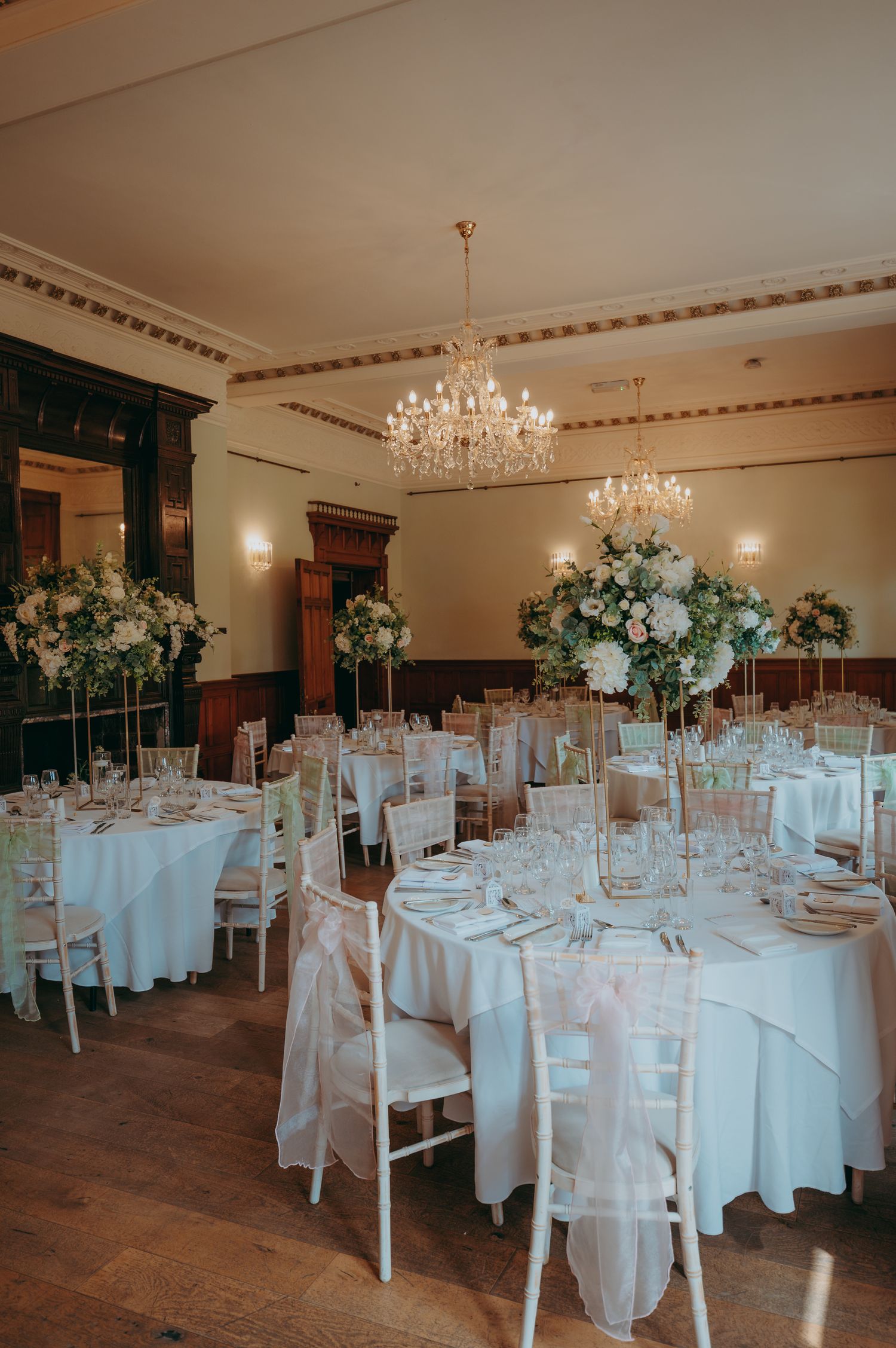 Elegant wedding reception hall with white round tables, chiavari chairs, tall floral centerpieces, and a crystal chandelier.