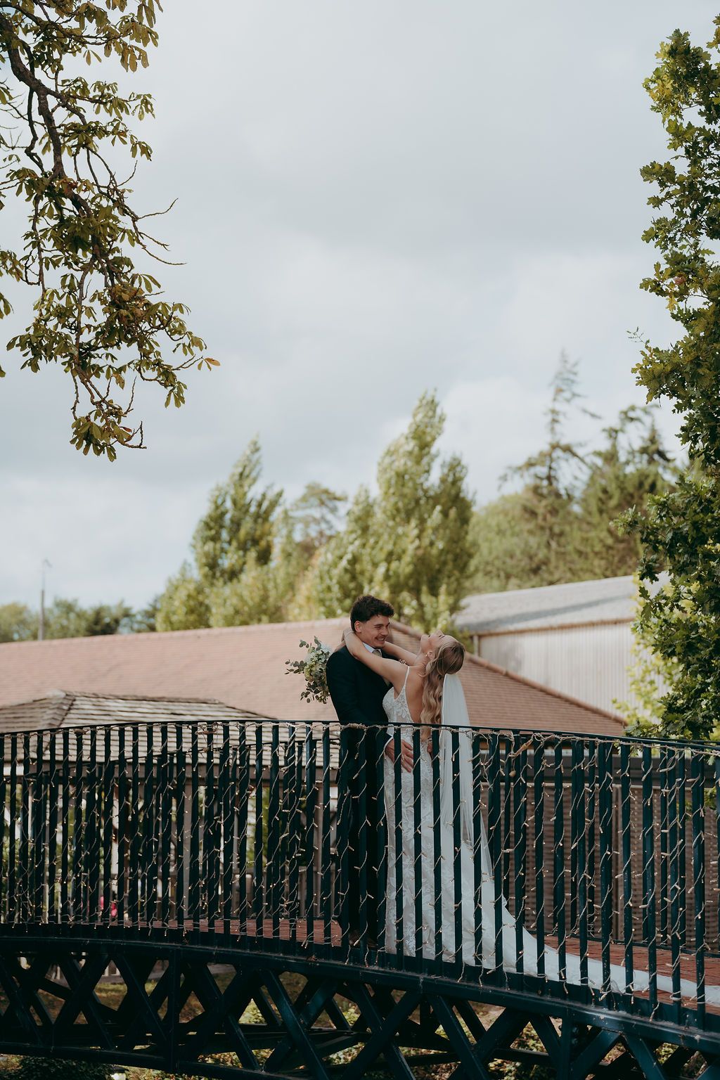 Bride in a long veil and groom in a dark suit embrace on an ornate black iron bridge surrounded by green trees.