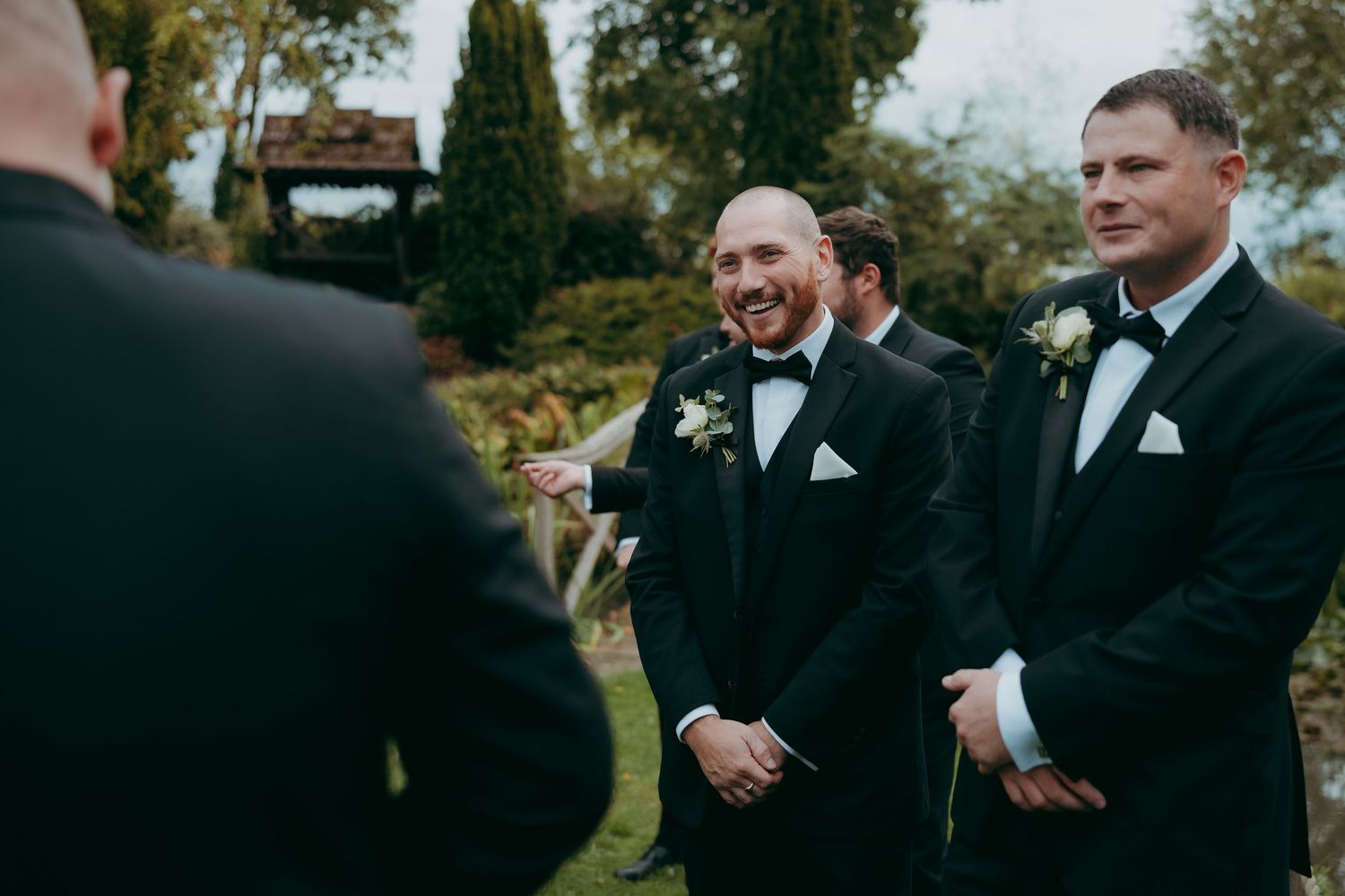 Groom in black tuxedo laughing with groomsmen wearing boutonnieres at outdoor wedding ceremony.