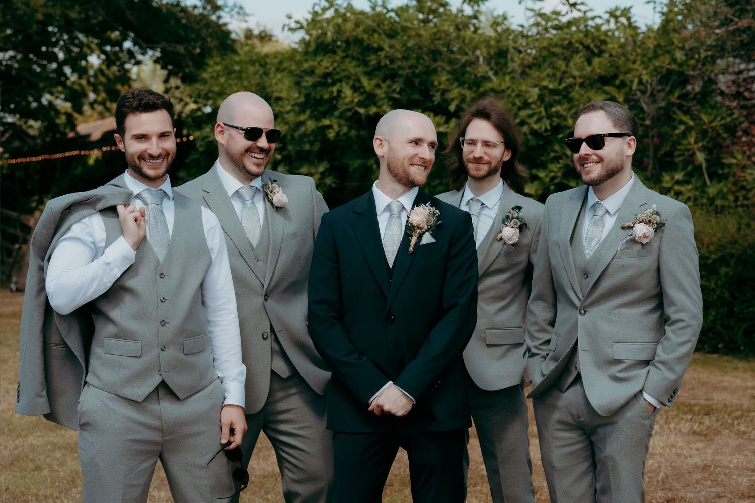 Groom in navy suit stands with four groomsmen in grey suits and boutonnieres, posing outdoors at a wedding.