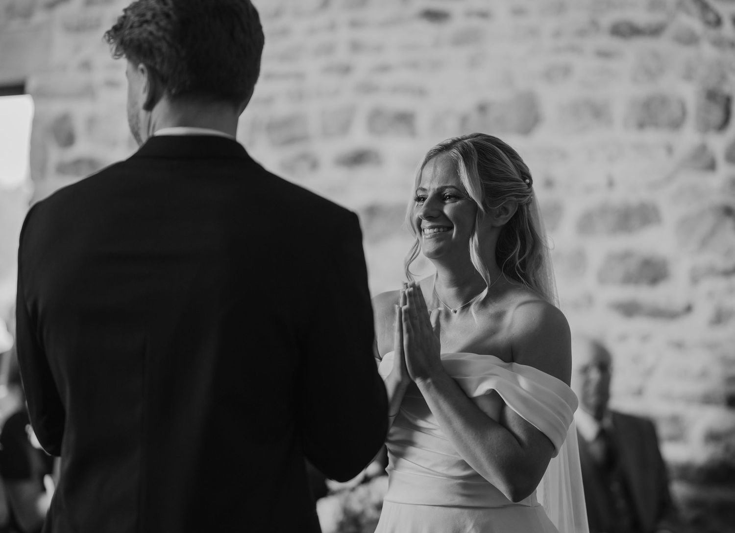 Black and white wedding photo of smiling bride in off-shoulder gown exchanging vows with groom at ceremony.