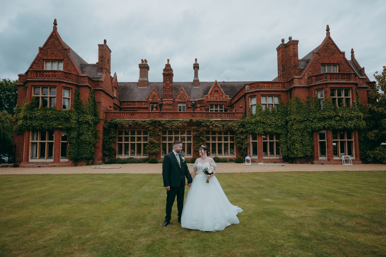 Bride and groom pose on lawn before grand red brick Victorian manor wedding venue on cloudy day.