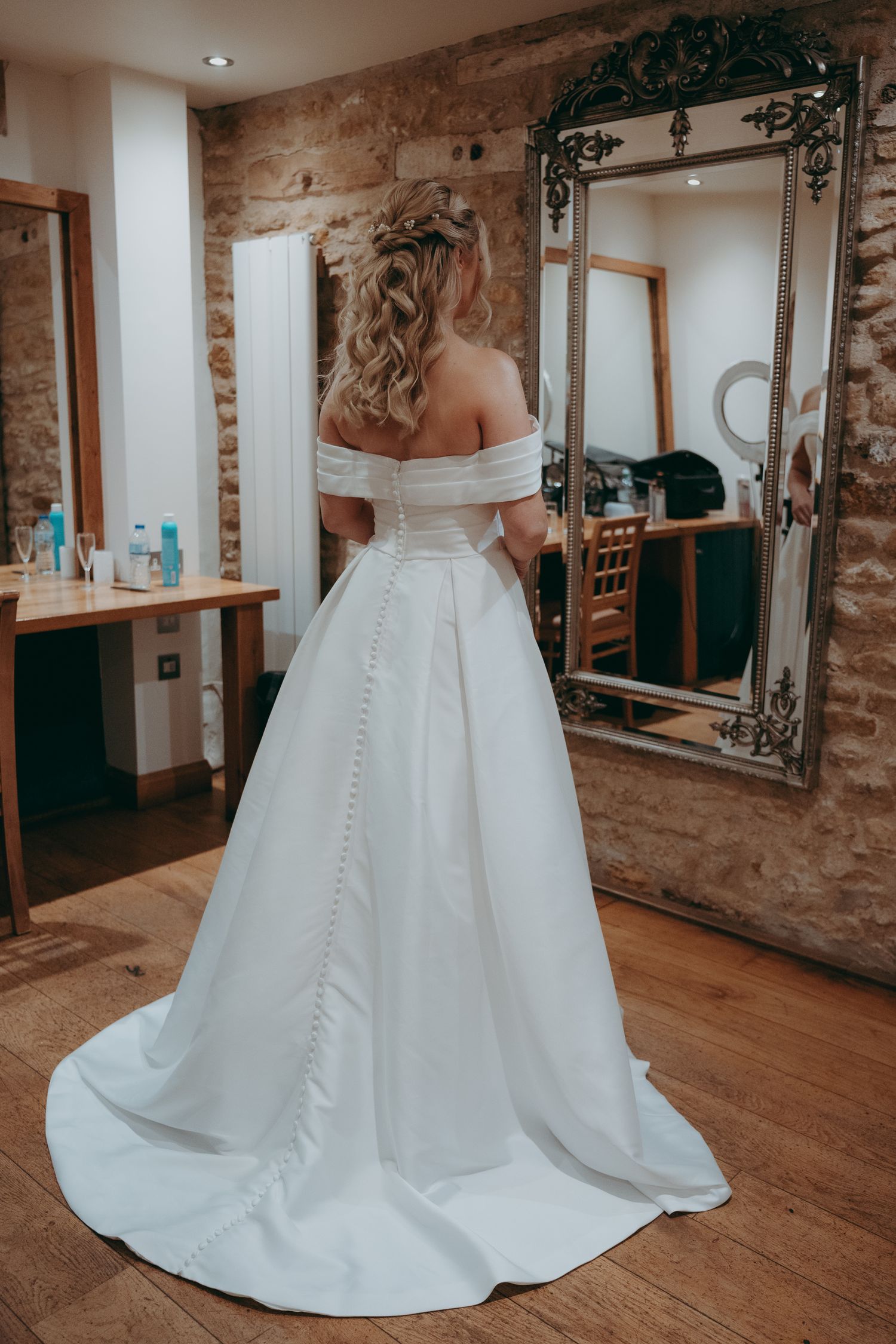 Bride in elegant off-shoulder white ball gown with button back detail, standing before ornate mirror in rustic venue.