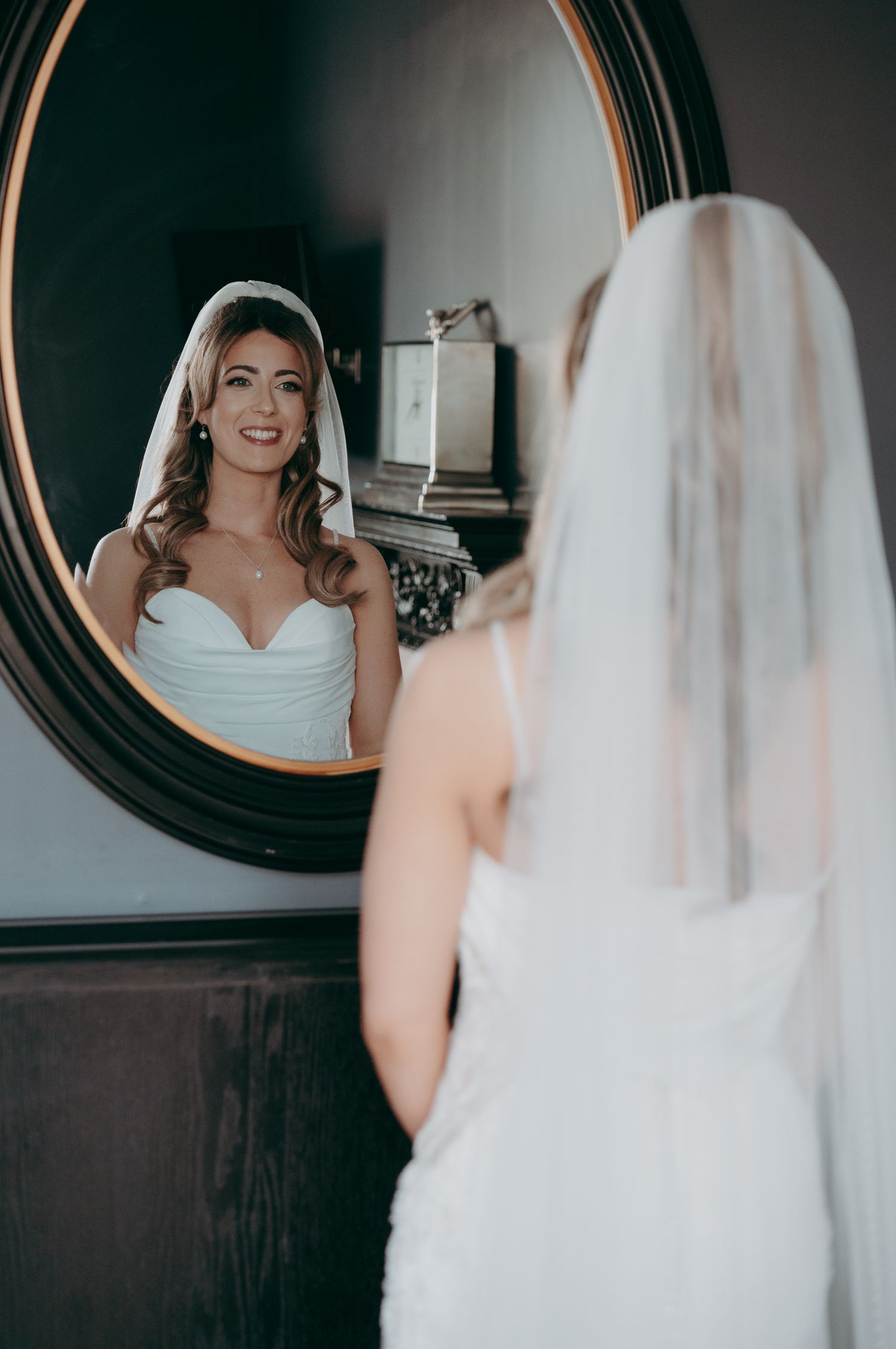 Bride in white strapless gown and veil admiring her reflection in an ornate round mirror on her wedding day.