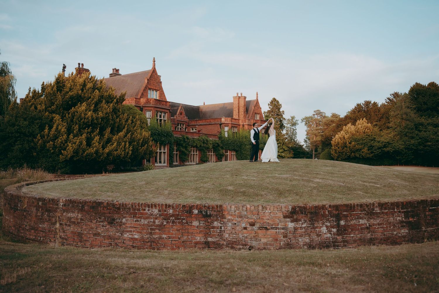 Bride and groom stand atop a grassy hill beside a grand red brick manor house surrounded by autumn trees.