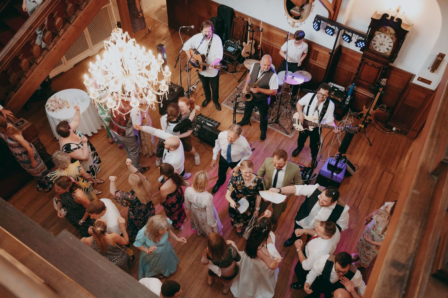 Aerial view of wedding reception with guests dancing on hardwood floor while live band performs in elegant venue.