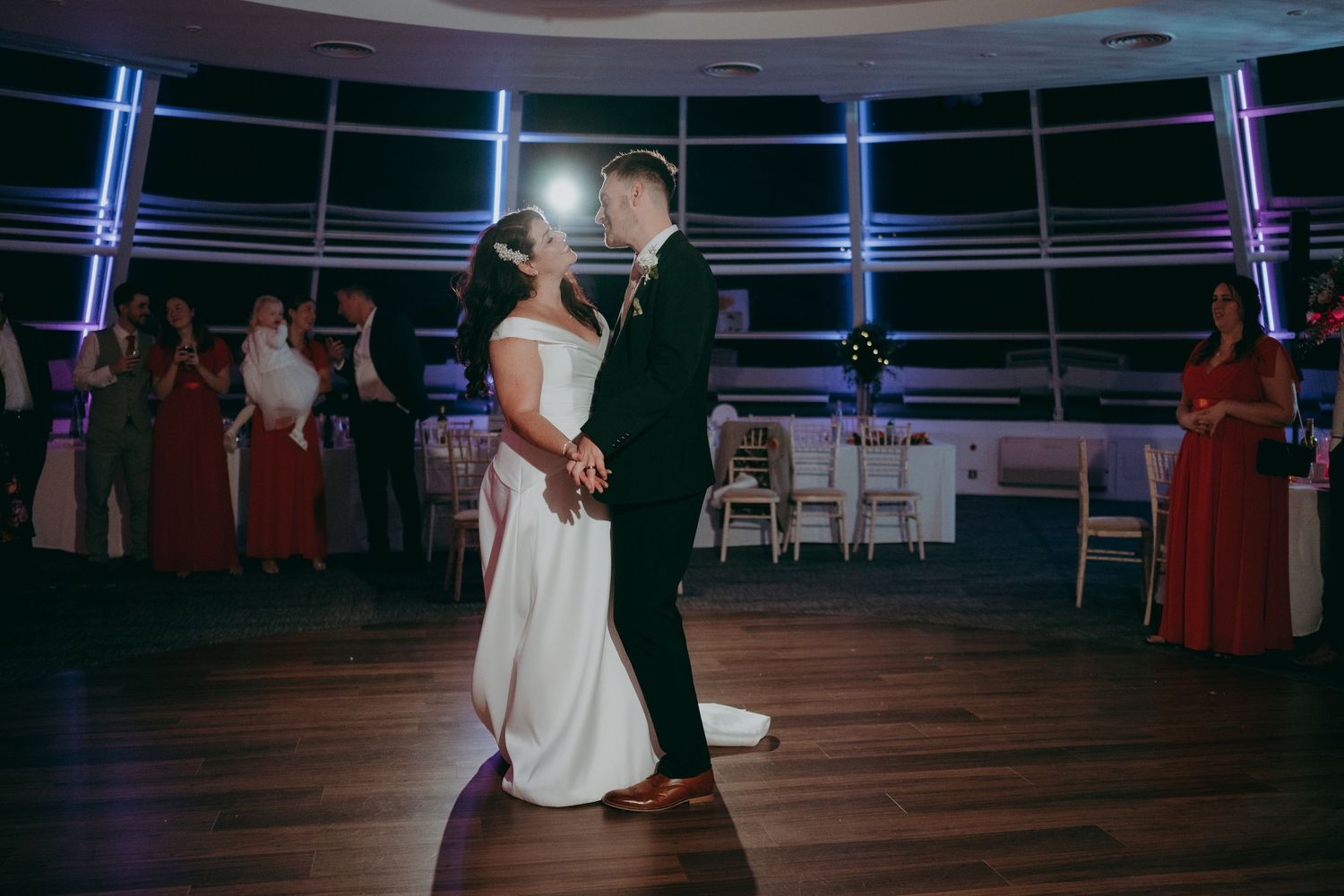 Bride and groom share their first dance at elegant wedding reception with blue lighting and guests watching in background.