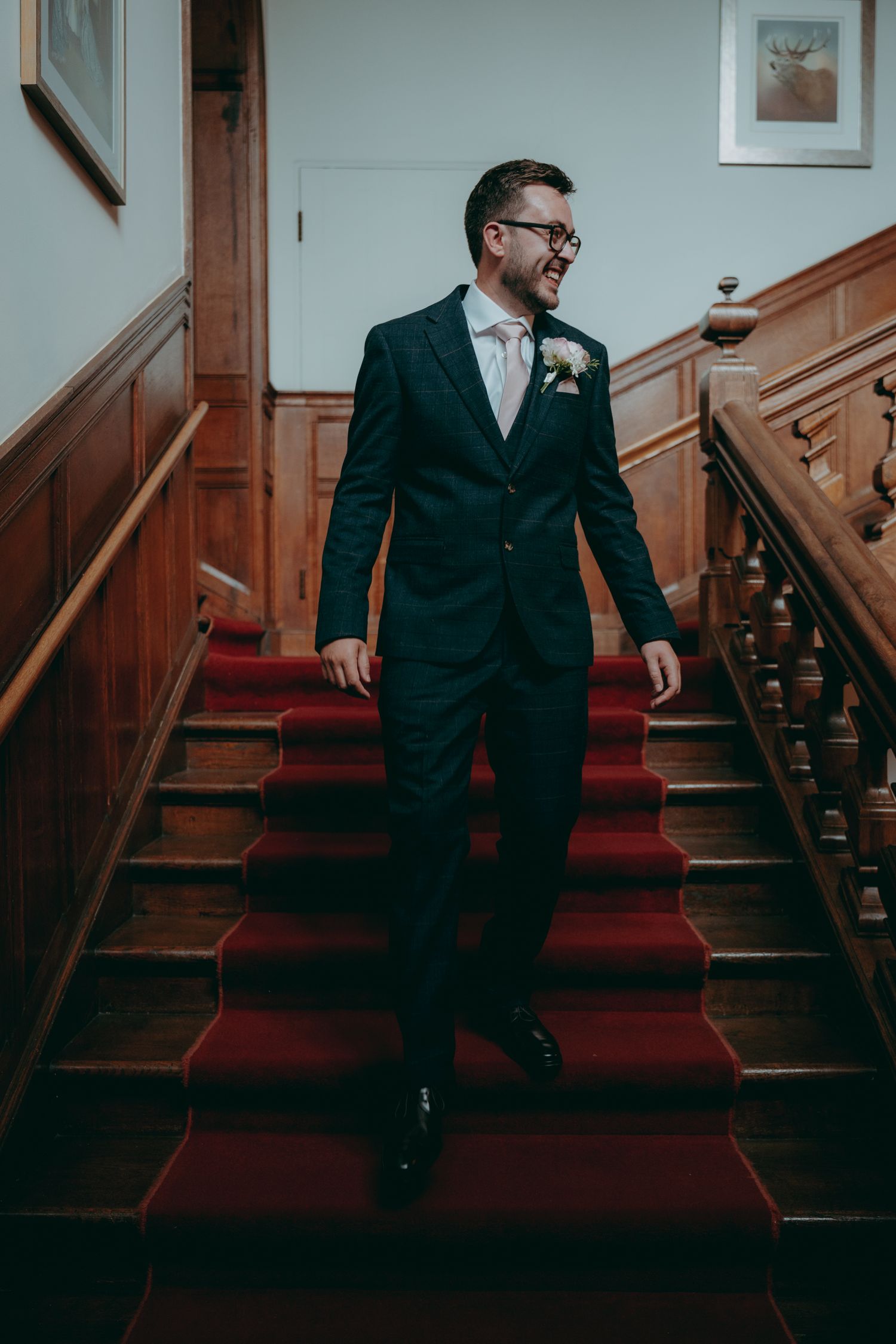 Smiling groom in navy suit with boutonniere descends red-carpeted wooden staircase in elegant venue.