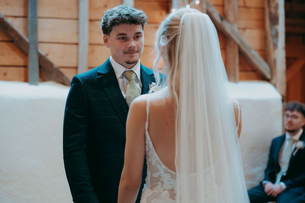 Groom in a dark suit smiles at his bride in a lace gown and long veil during a rustic barn wedding ceremony with a guest looking on.