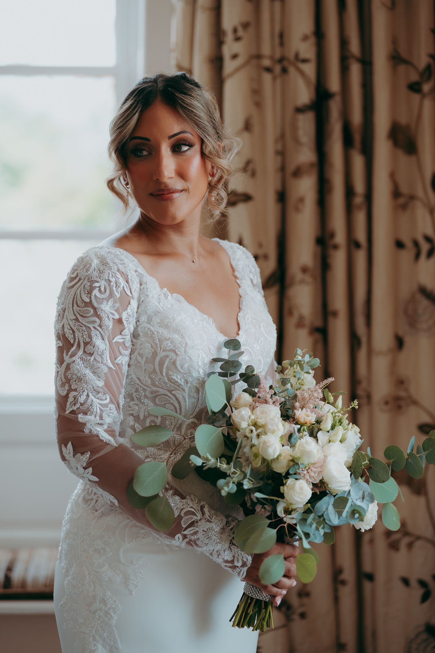 Bride in lace long-sleeve wedding dress holds white and blush bouquet with eucalyptus near a sunlit window.