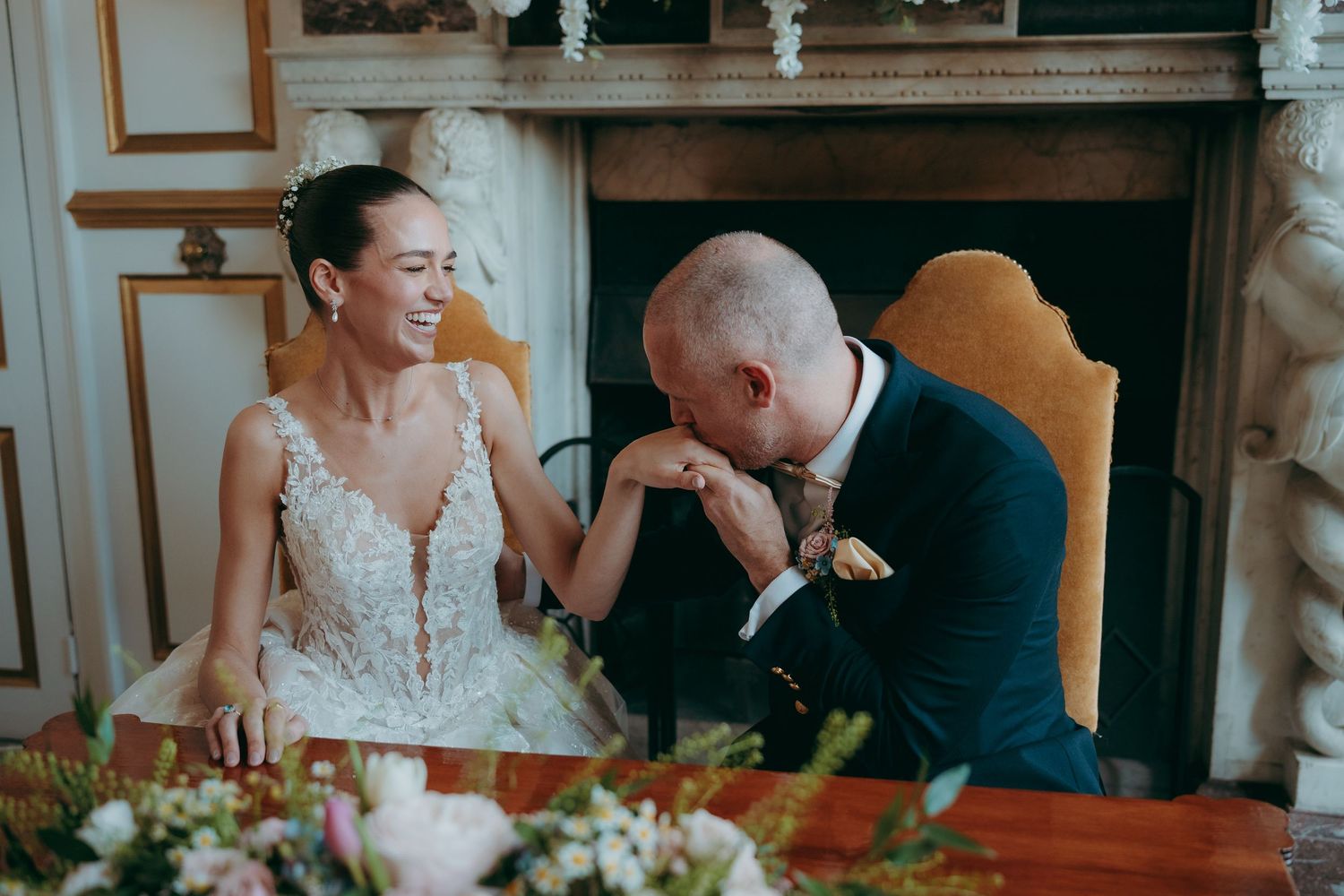 Bride and groom share a joyful moment at wedding table, groom kissing bride's hand near floral centerpiece.