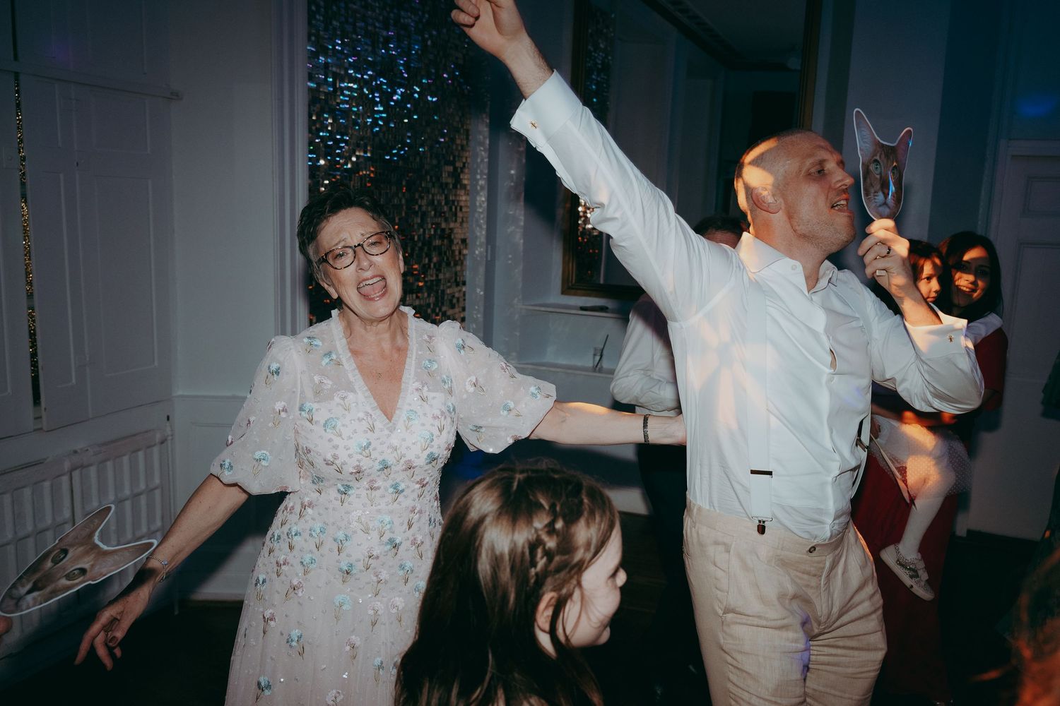 Couple dancing joyfully at a wedding reception, woman in floral dress and man in white shirt raising arms.