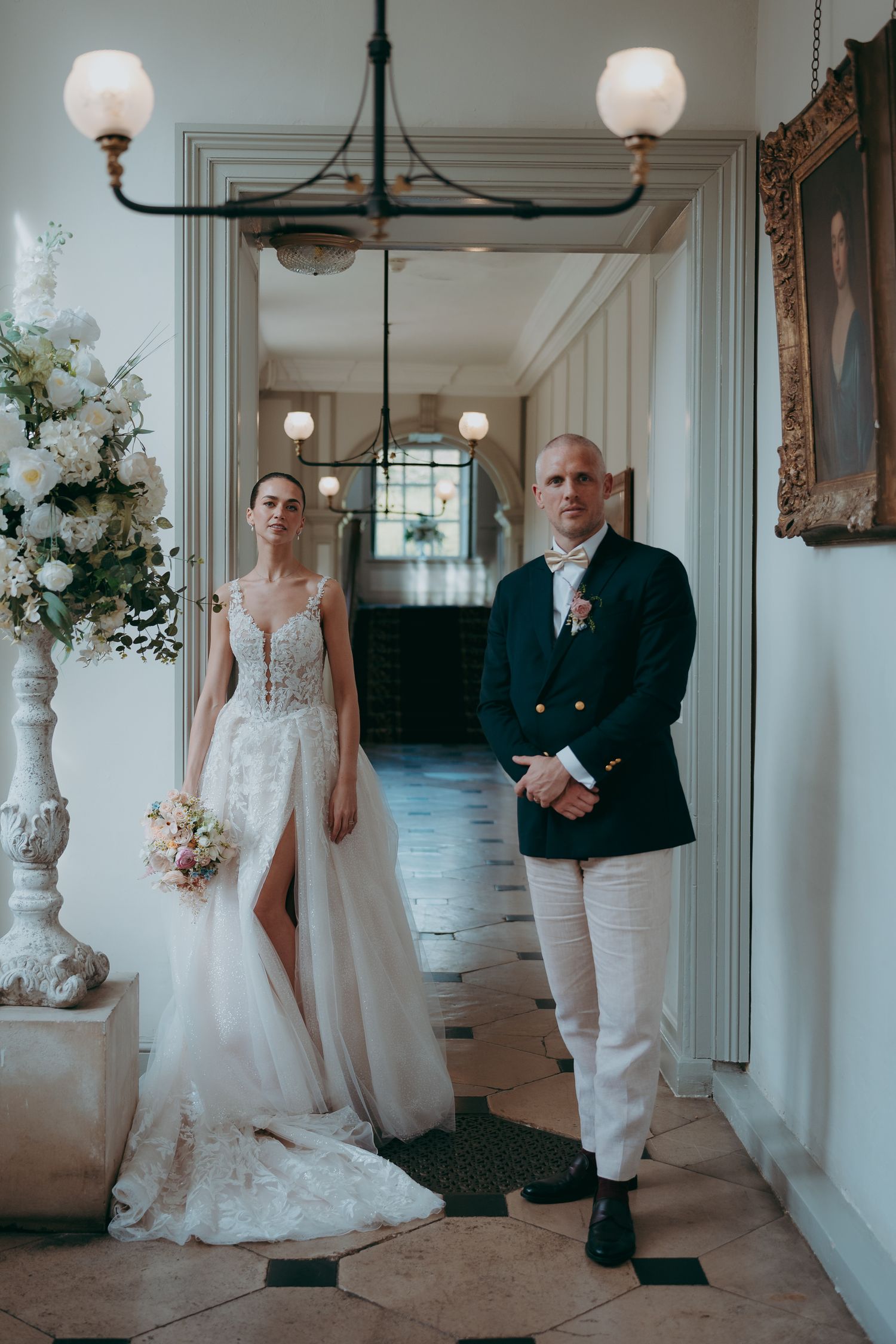 Bride in lace gown holding bouquet stands with groom in navy blazer and white pants in elegant venue hallway.