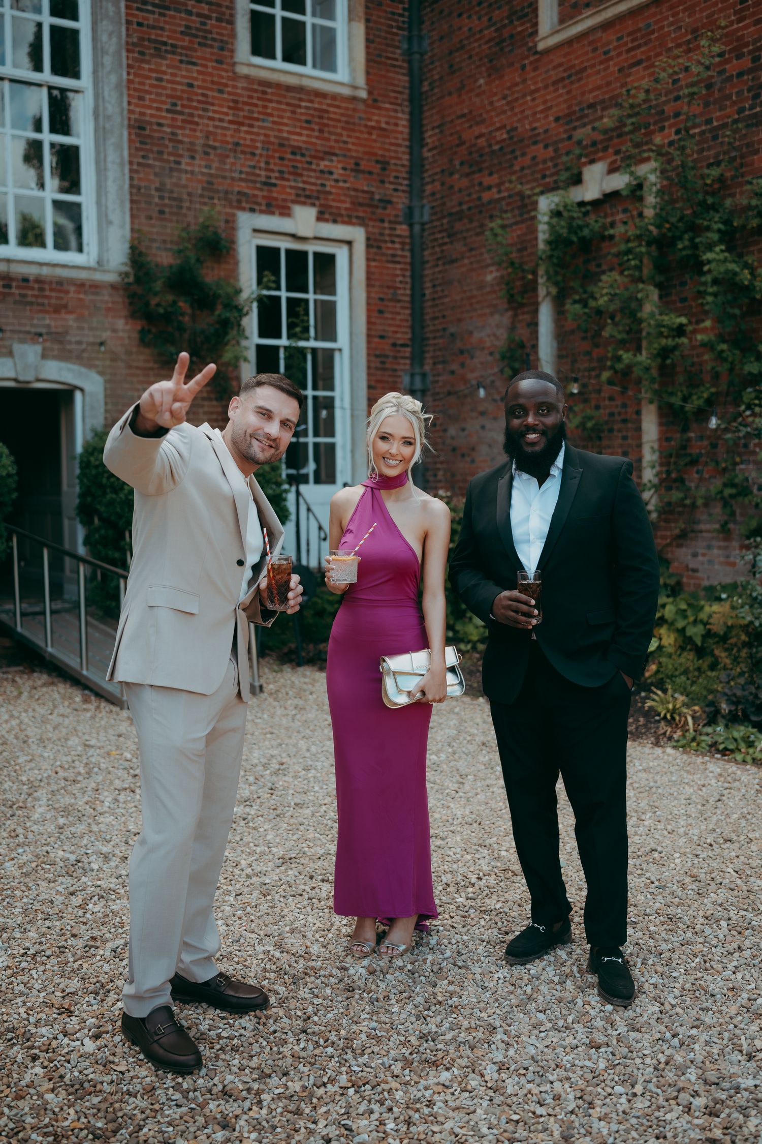 Three wedding guests posing outdoors at a brick manor, woman in magenta dress flanked by men in suits.