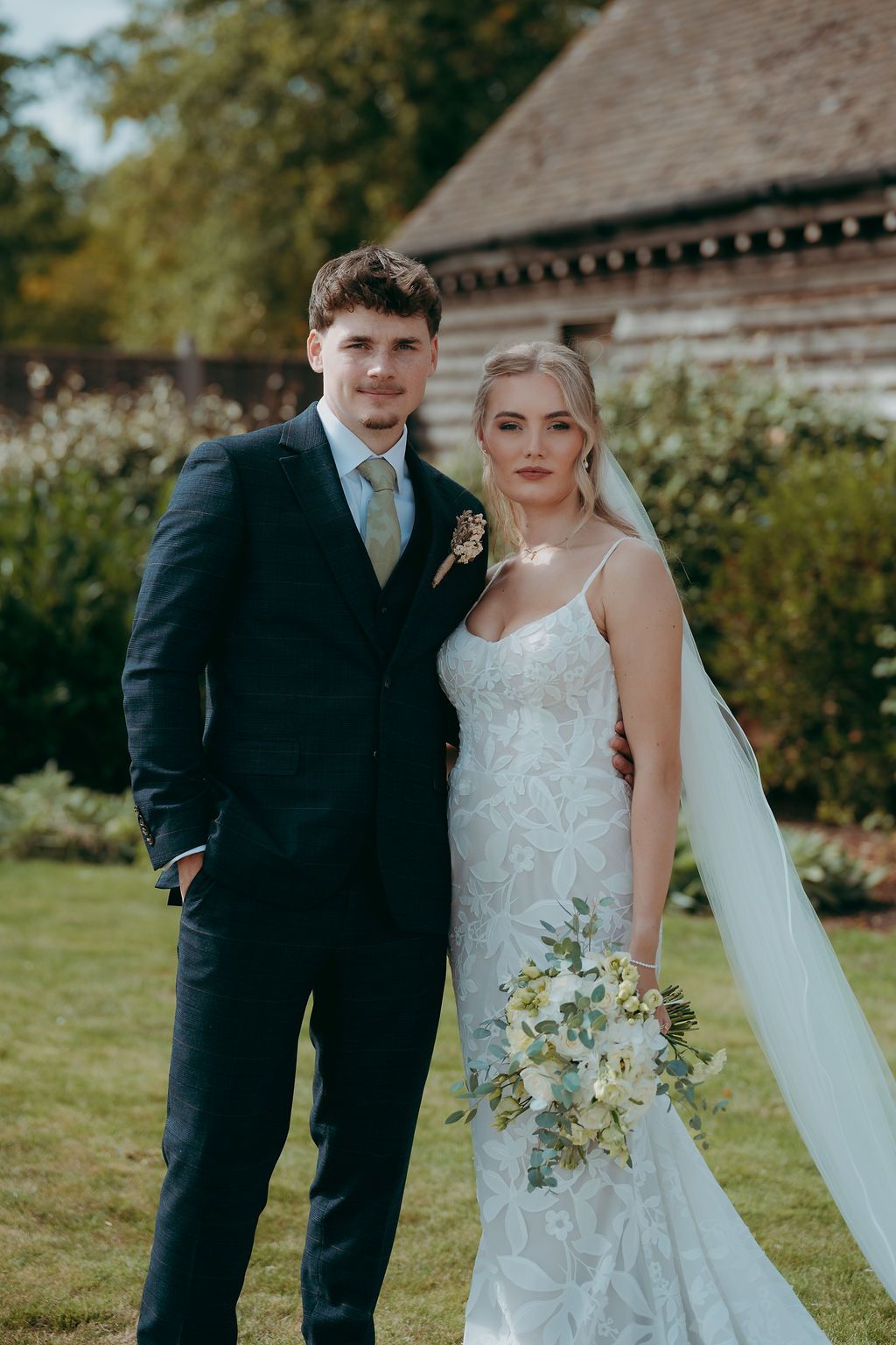Bride in a lace wedding gown and veil holding a floral bouquet stands beside a groom in a navy three-piece suit in a sunny garden.