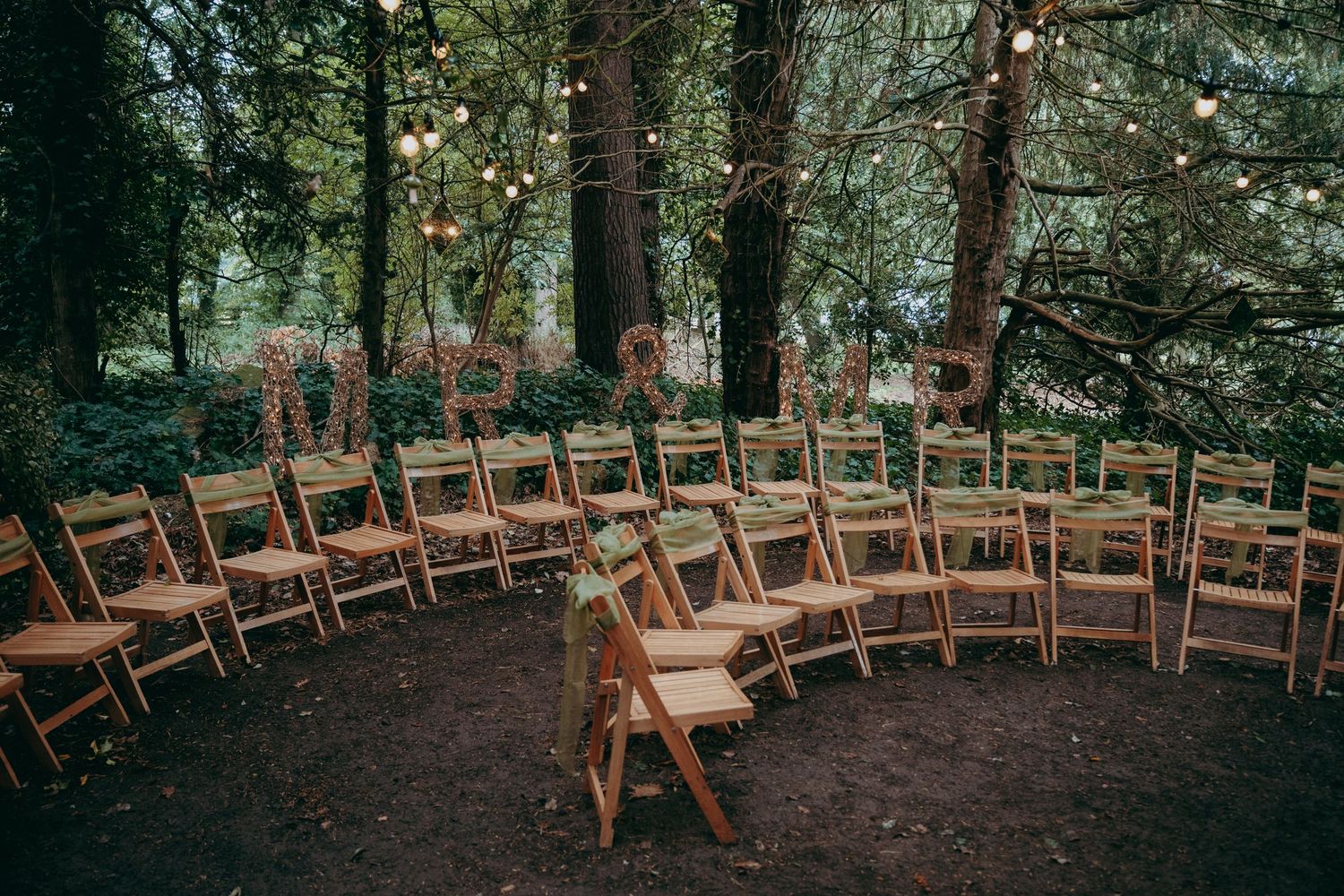 Wooden folding chairs arranged in a woodland wedding ceremony space with fairy lights and rustic letter decorations.