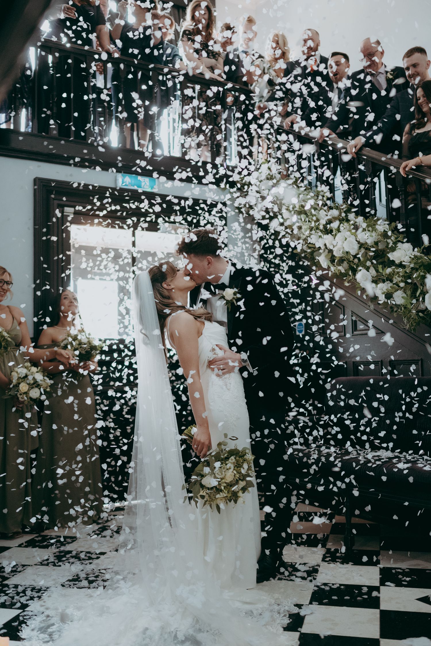 Bride and groom share a kiss as white confetti rains down during their wedding ceremony exit.