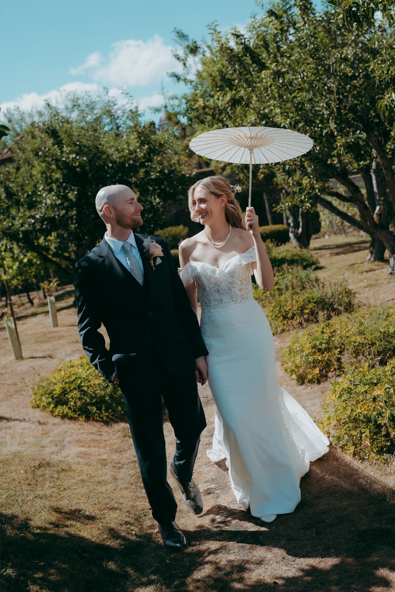 Bride and groom walking outdoors on sunny day, bride in lace gown holding white parasol, groom in dark suit.