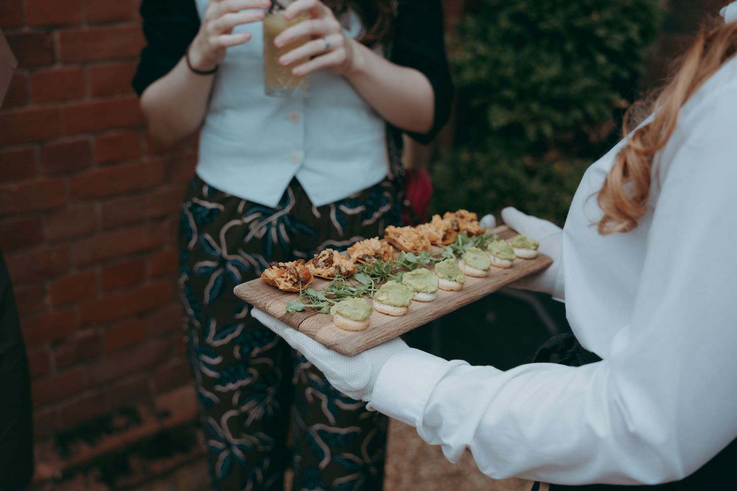 Server holding a wooden charcuterie board with appetizers at an outdoor catering event near a brick wall.