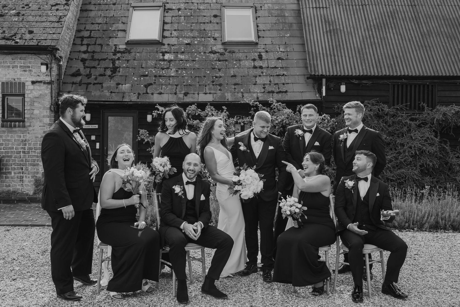 Black and white wedding party photo outside rustic barn venue with bride, groom, and elegantly dressed guests.