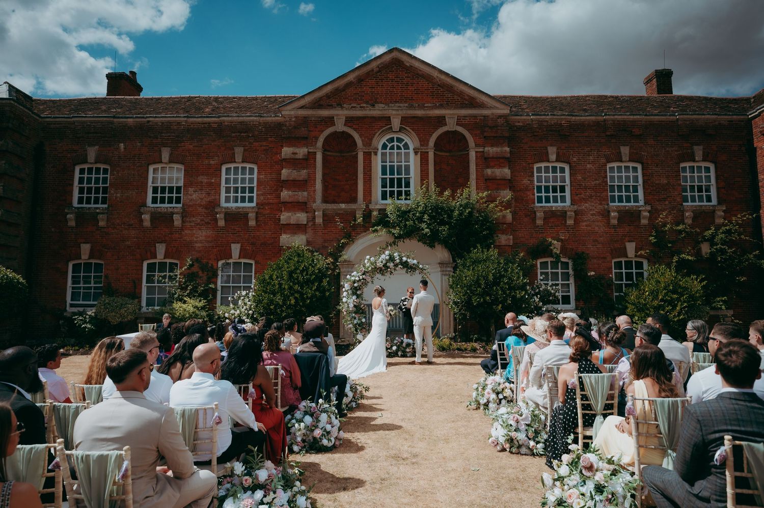 Outdoor wedding ceremony at a grand red brick Georgian manor house with floral arch altar and seated guests on a sunny day.