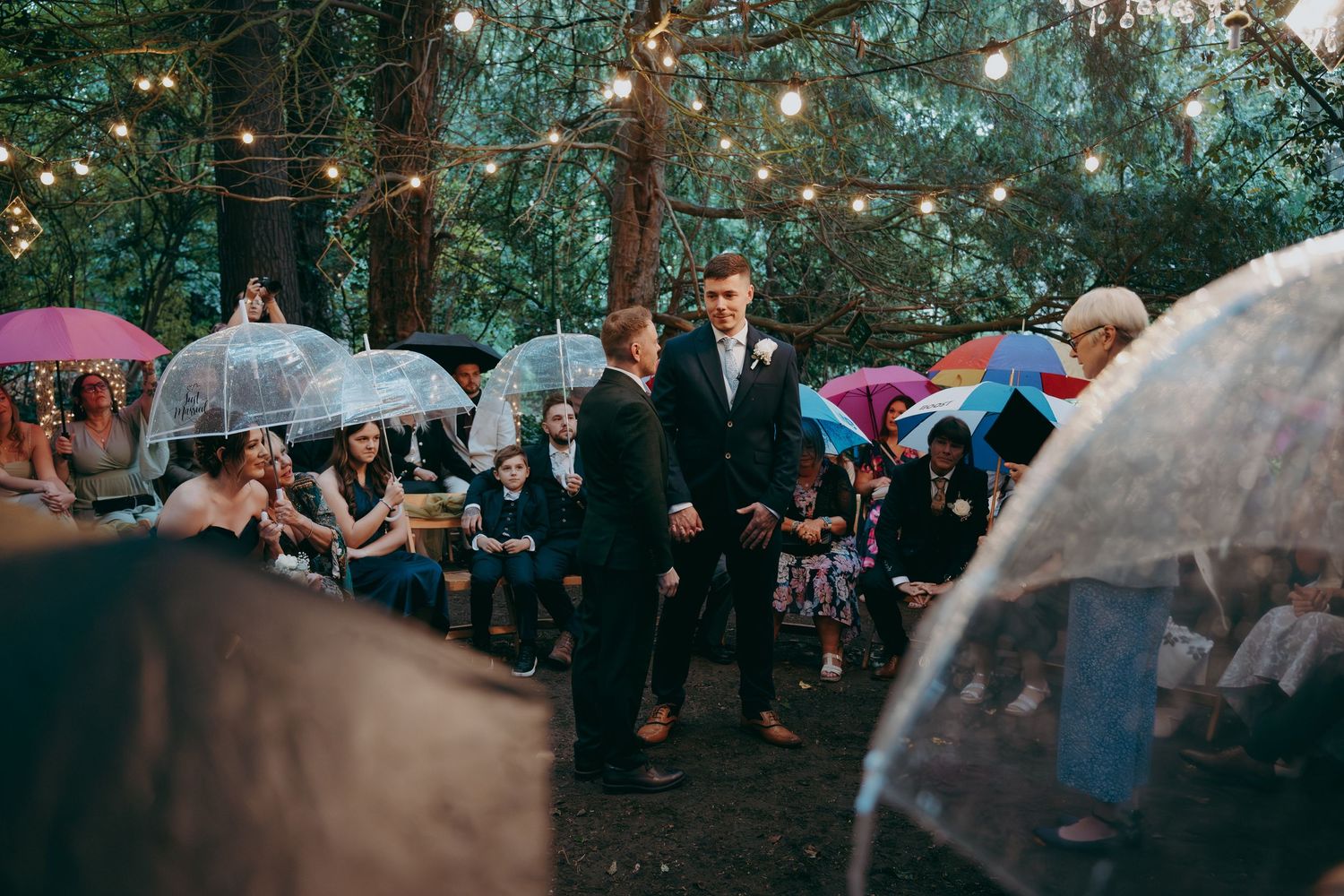 Two grooms exchange vows at a rainy outdoor wedding ceremony surrounded by guests holding clear umbrellas with string lights.