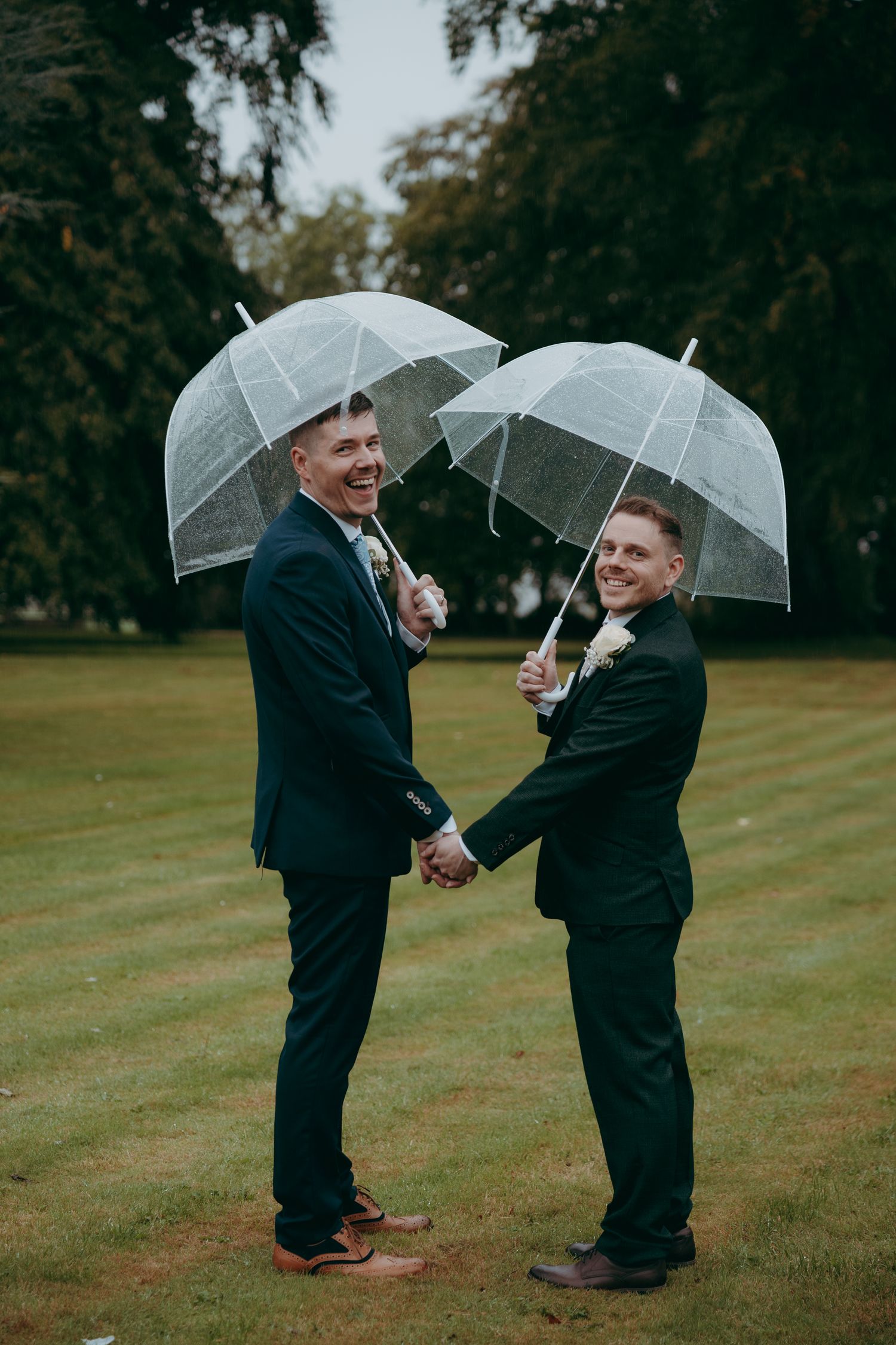 Two grooms in navy suits hold clear umbrellas and hands, smiling joyfully on a lush green lawn on their wedding day.