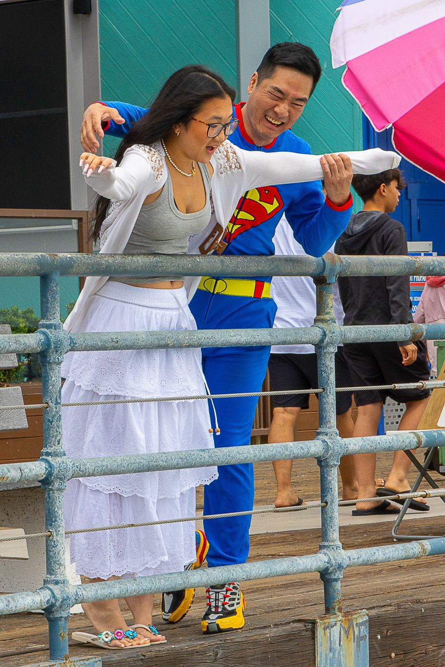 Man in Superman costume poses with a woman on a boardwalk, laughing and spreading his arms wide.