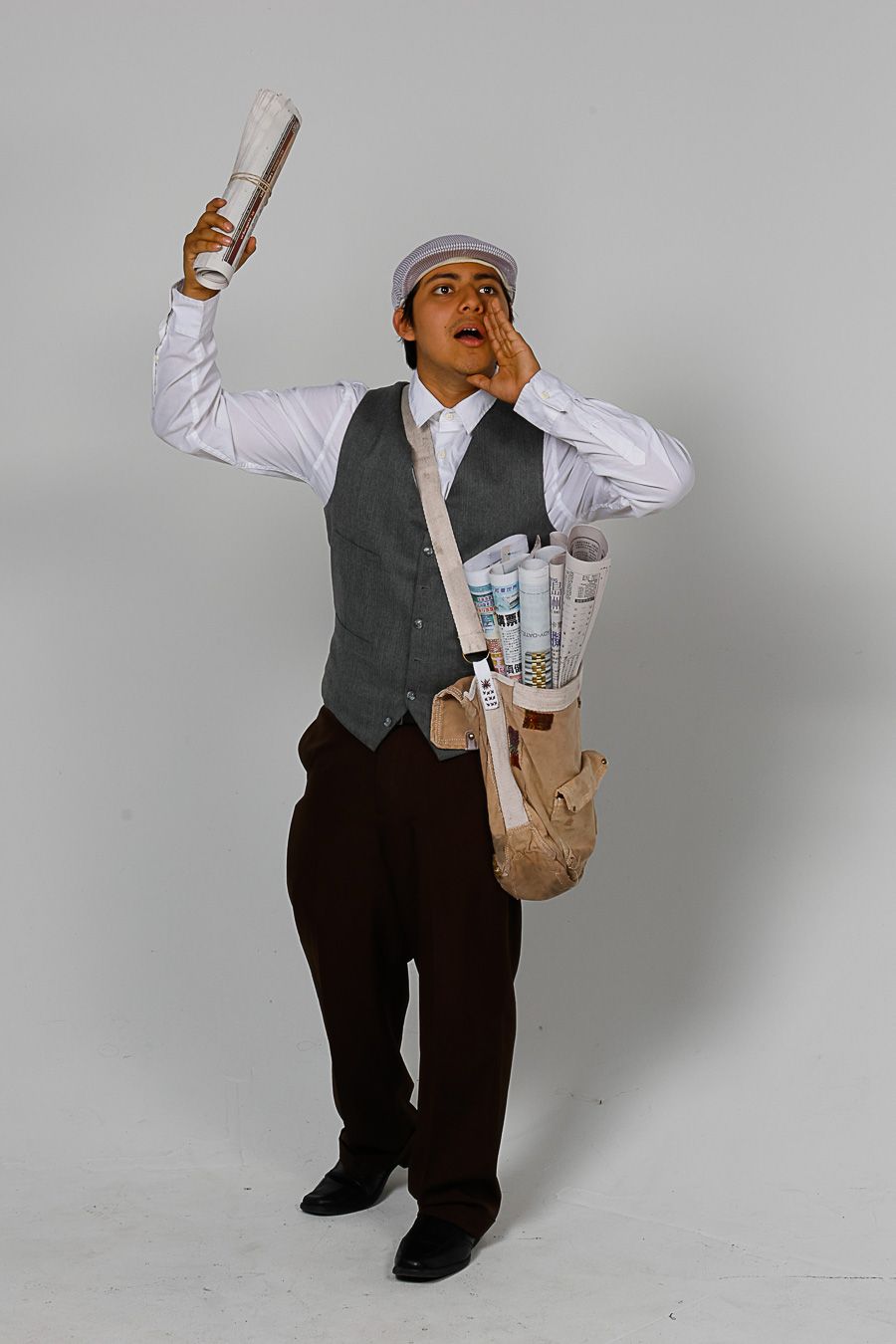A newsboy in vest and cap shouts while holding a rolled newspaper, with a bag full of papers over his shoulder.