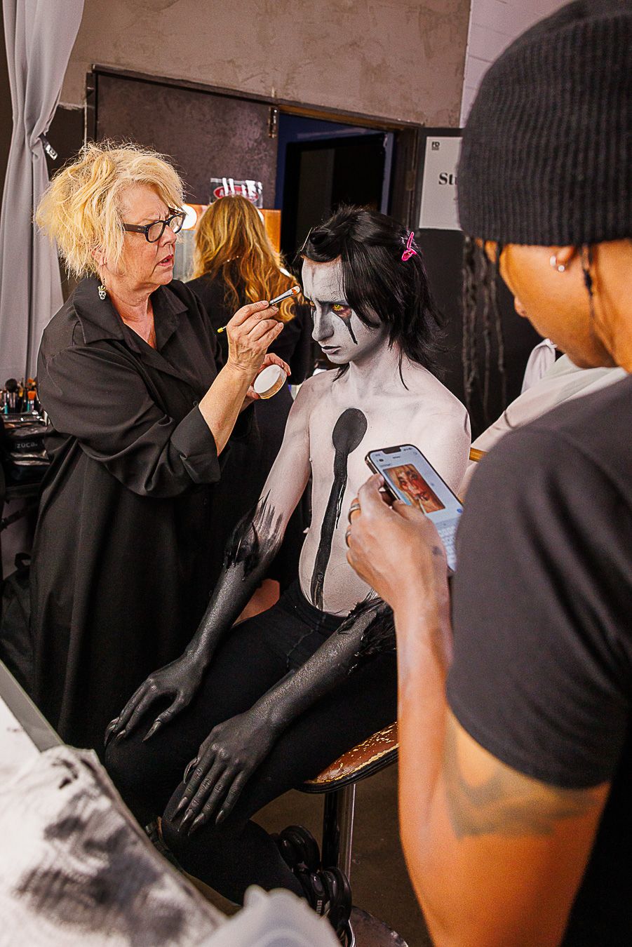Makeup artist applying black and white body paint to a model in a backstage studio setting.