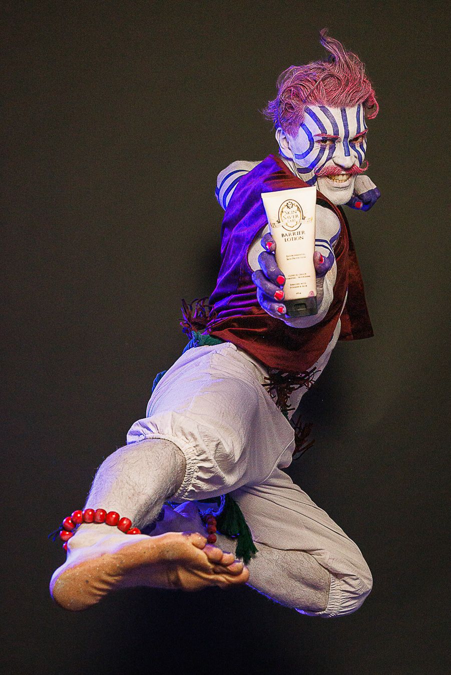 Performer in white face paint and pink hair jumps mid-air holding a product tube, wearing colorful costume.