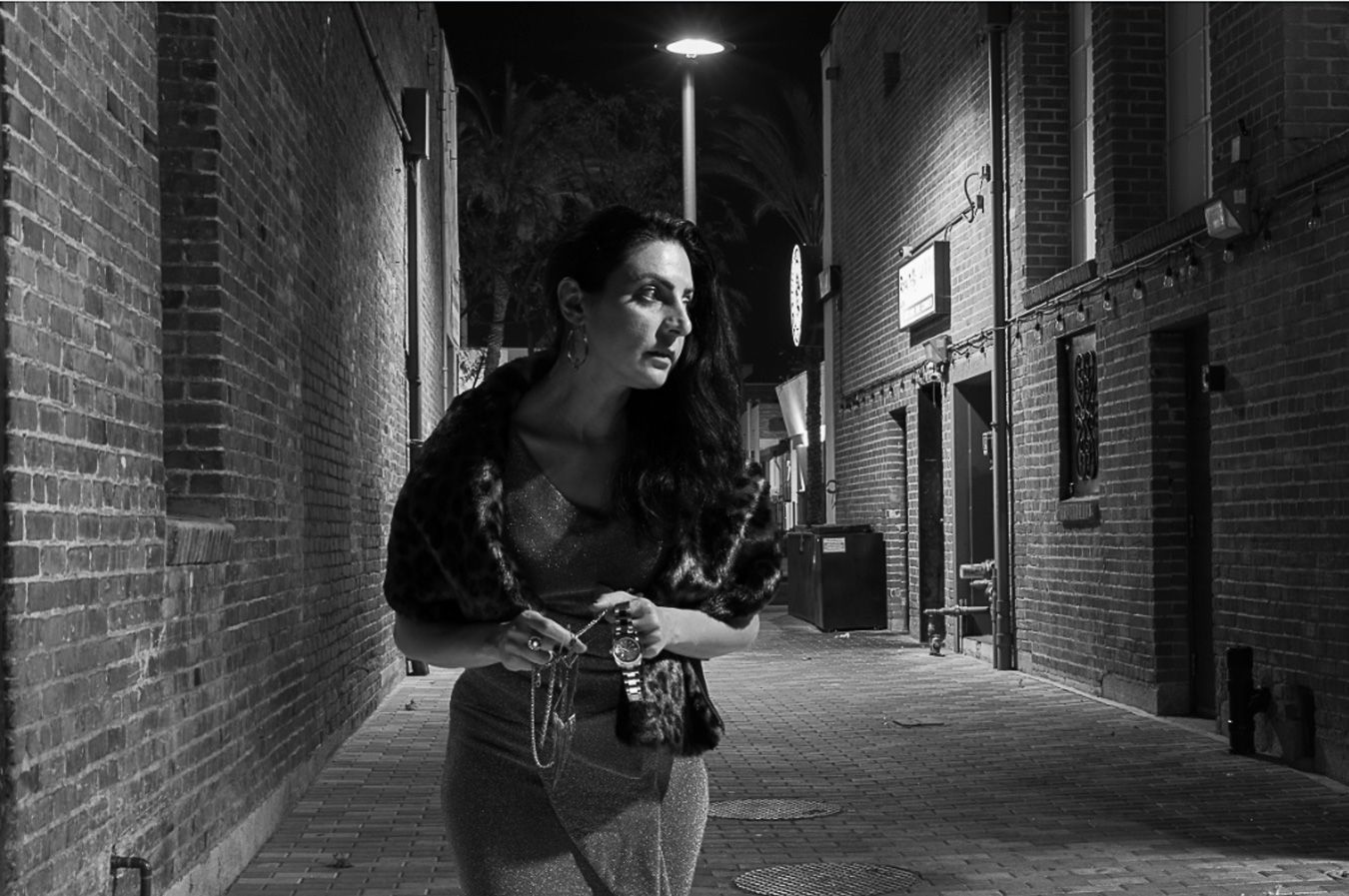 Black and white photo of a woman holding a camera in a dimly lit urban alley at night.