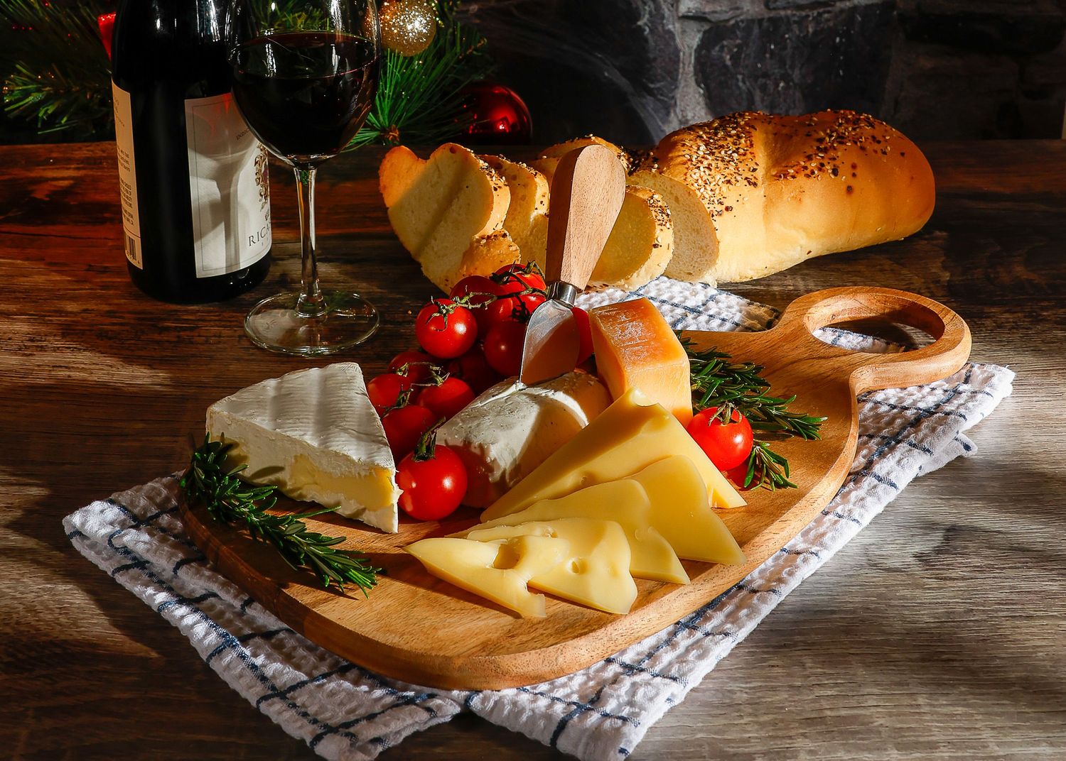 Rustic cheese board with brie, tomatoes, rosemary, bread, and red wine glasses on wooden table.