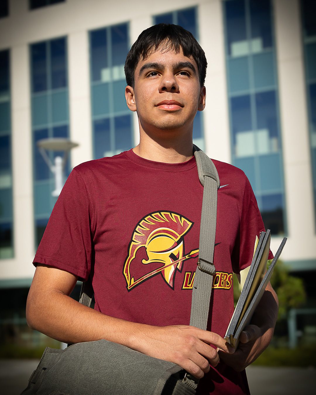 Young male student in maroon Lancers t-shirt holding books outdoors near modern building.