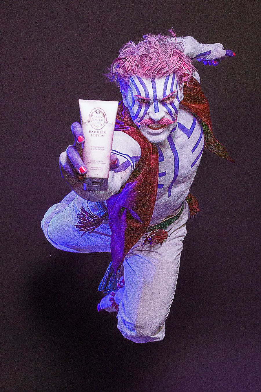 Performer in white body paint with blue tribal stripes and pink hair holds beauty product tube toward camera.