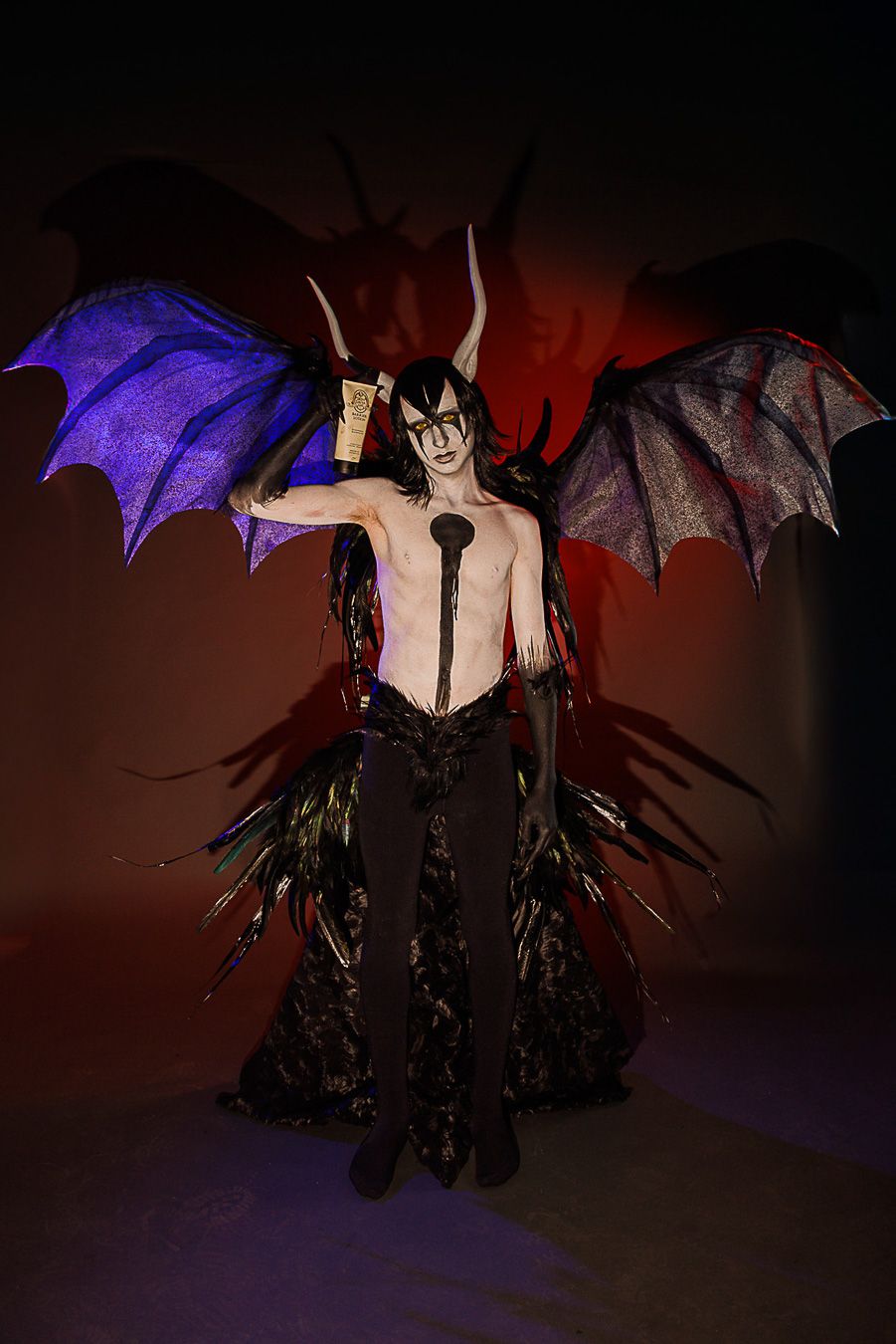 A performer in dramatic devil costume with large bat wings, horns, and body paint poses on a dark stage.