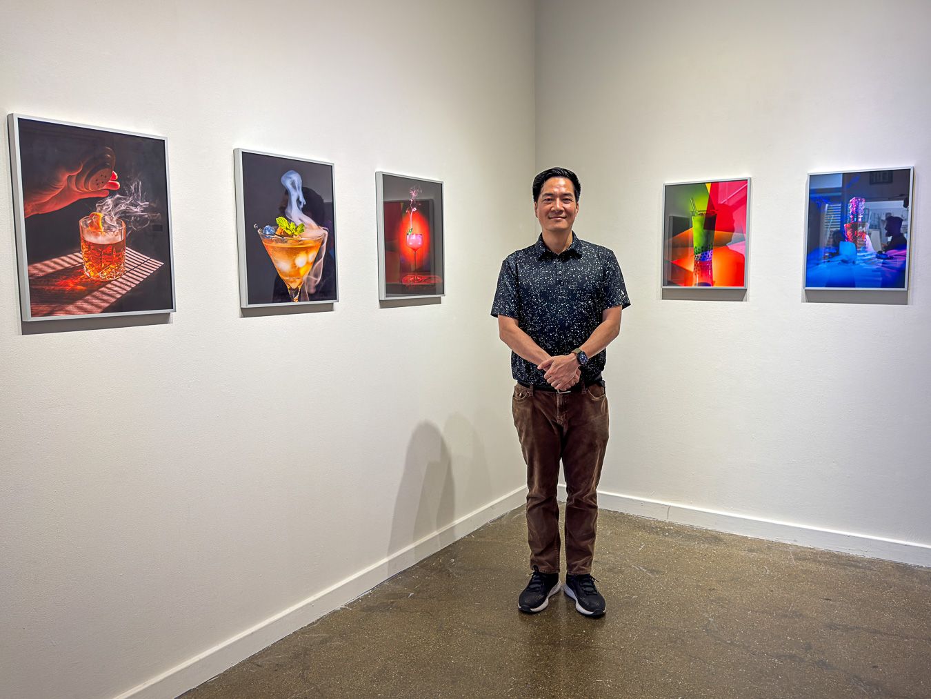 Man standing proudly in front of colorful food and abstract photography artwork displayed on white gallery walls.
