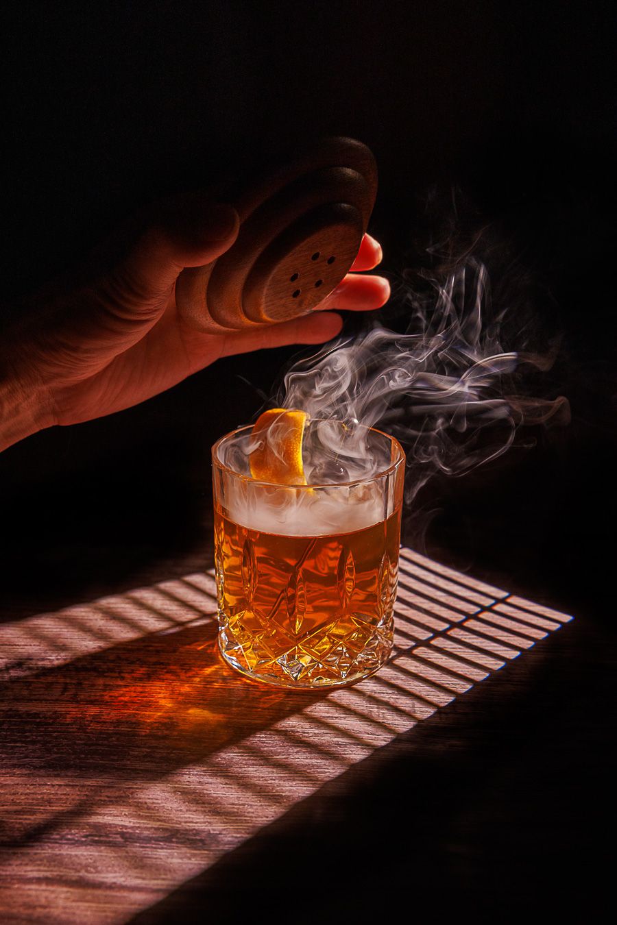 A smoky smashed Old Fashioned cocktail in a crystal glass with an orange peel, lit dramatically on a wooden surface.