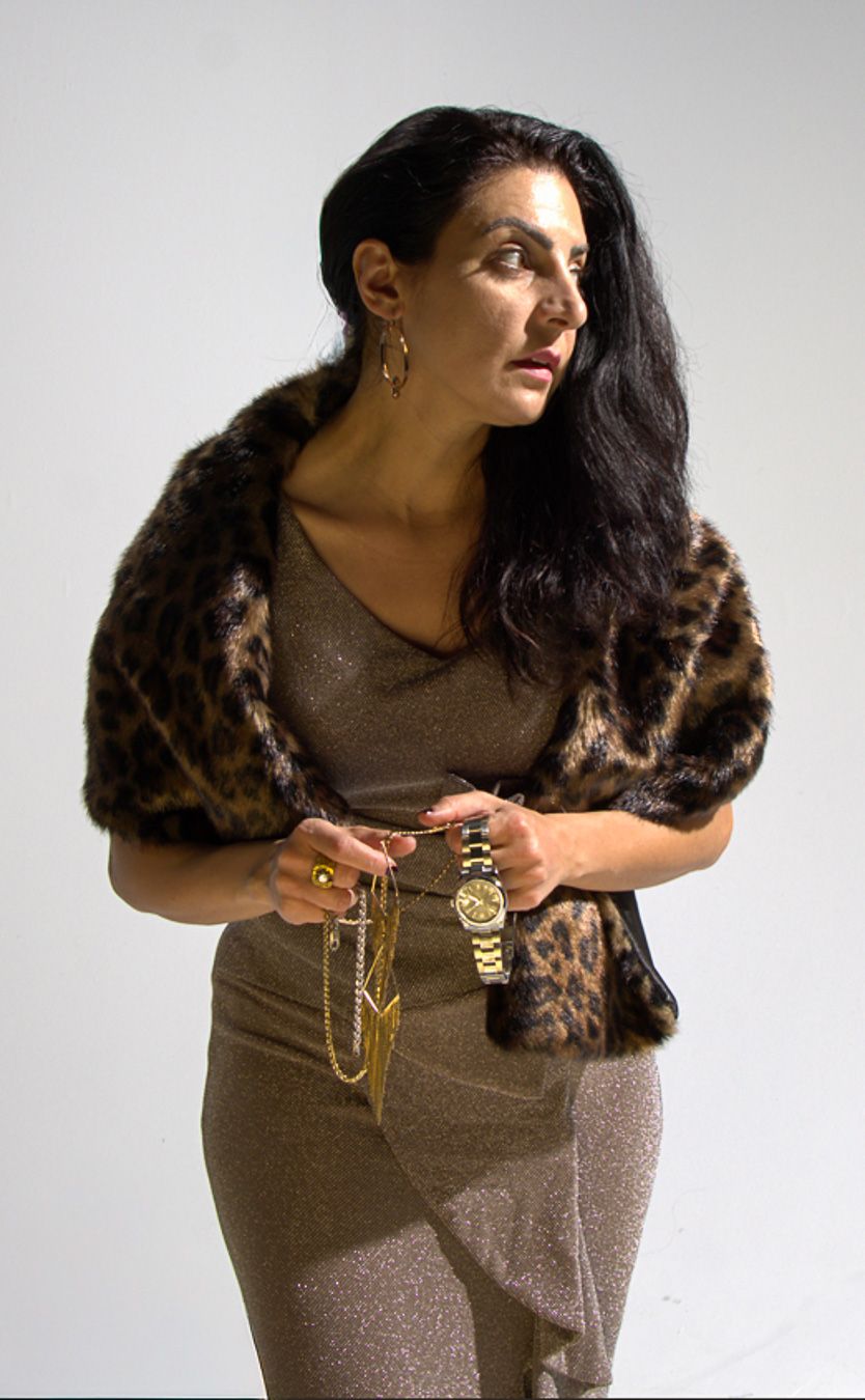 Woman in leopard fur stole and gold dress holding jewelry, wearing a Rolex watch, posing against white background.