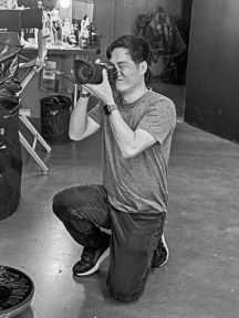 Black and white photo of man kneeling while taking photos with DSLR camera in studio.