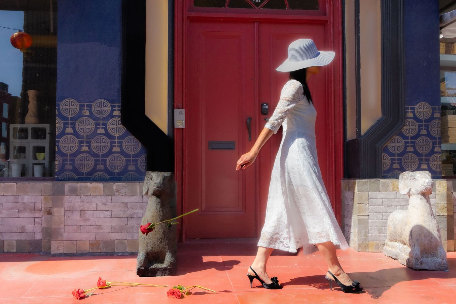 Woman in white dress and hat walks past stone lion statues on a red porch near a pink door.