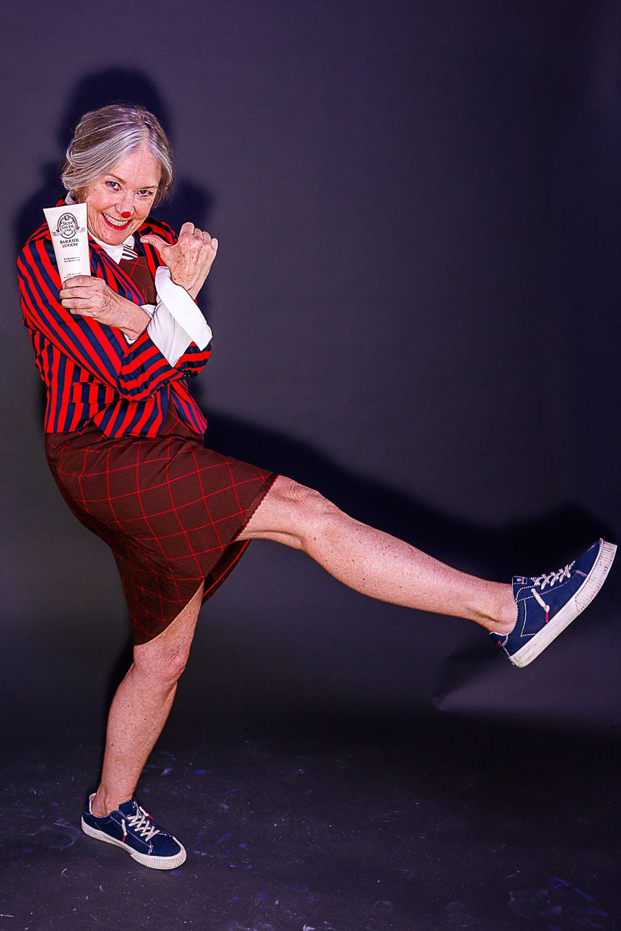 Energetic older woman in red striped blazer and plaid skirt kicks leg up while holding a white product tube.