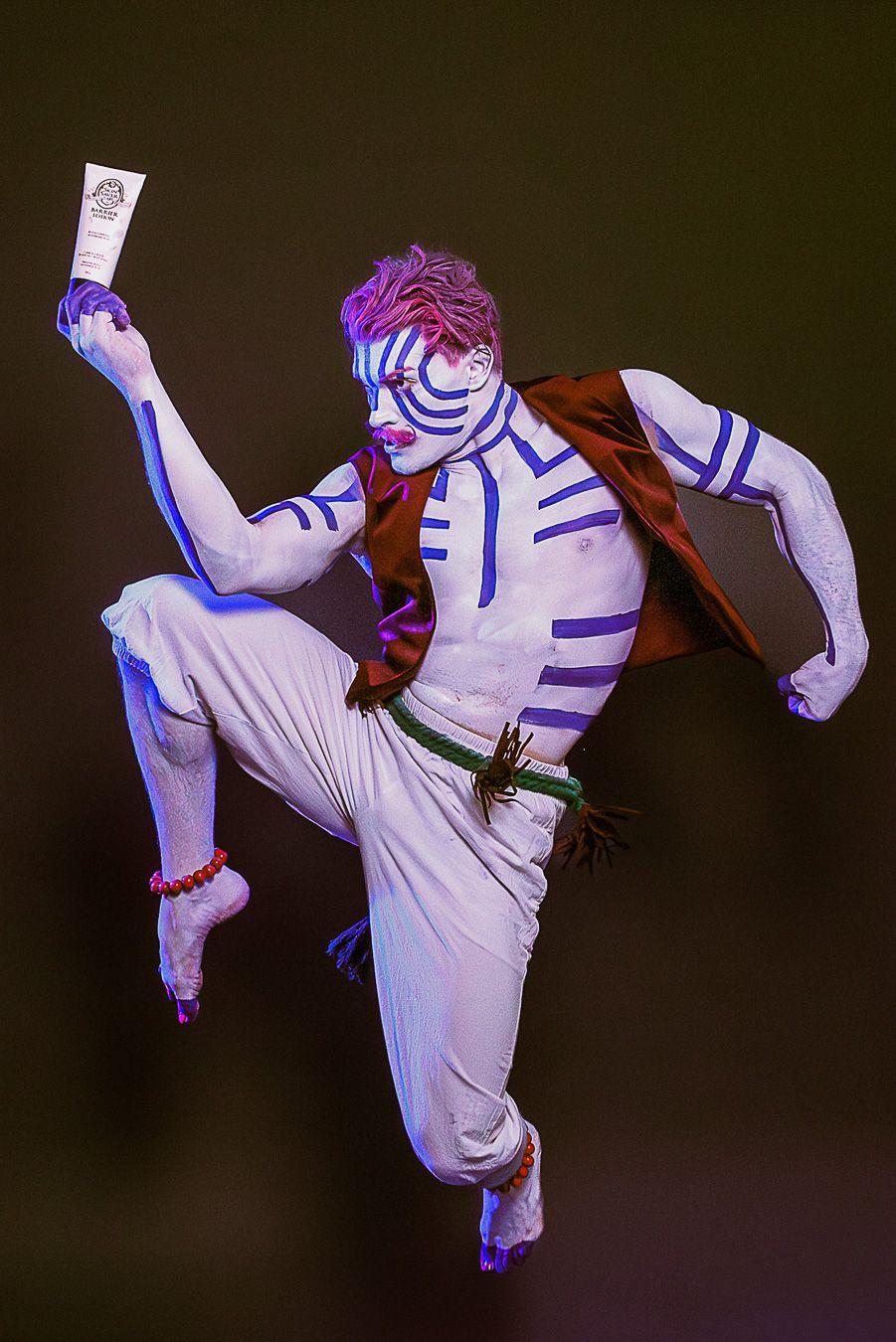 Performer in white body paint with blue stripes, dynamic pose, holding a tube, wearing white pants and brown vest.