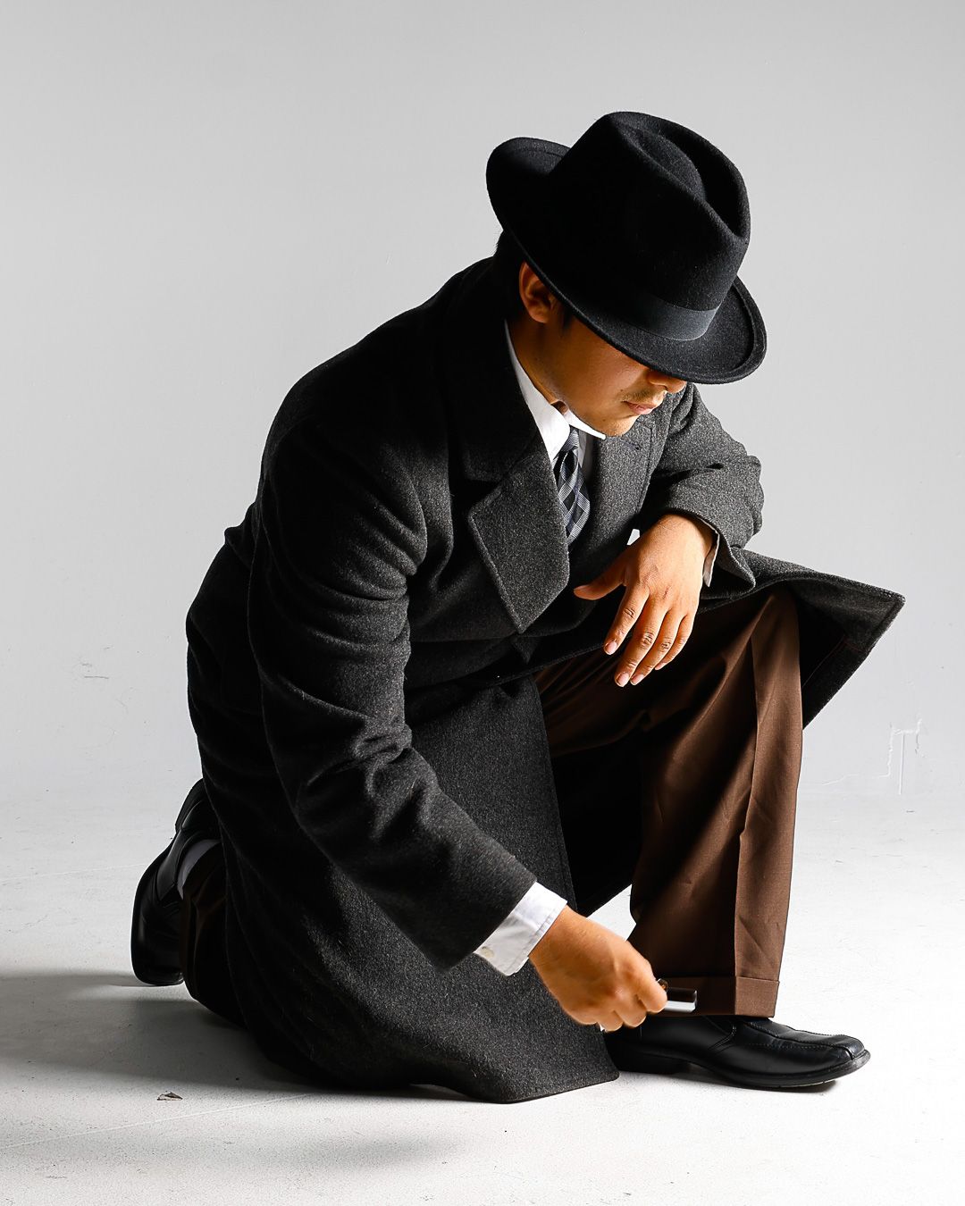 Man in black fedora hat and overcoat kneeling on floor in classic noir detective style pose.