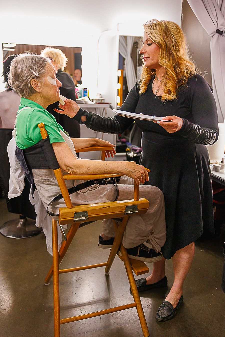 A makeup artist applies cosmetics to an elderly woman seated in a director's chair in a professional dressing room.