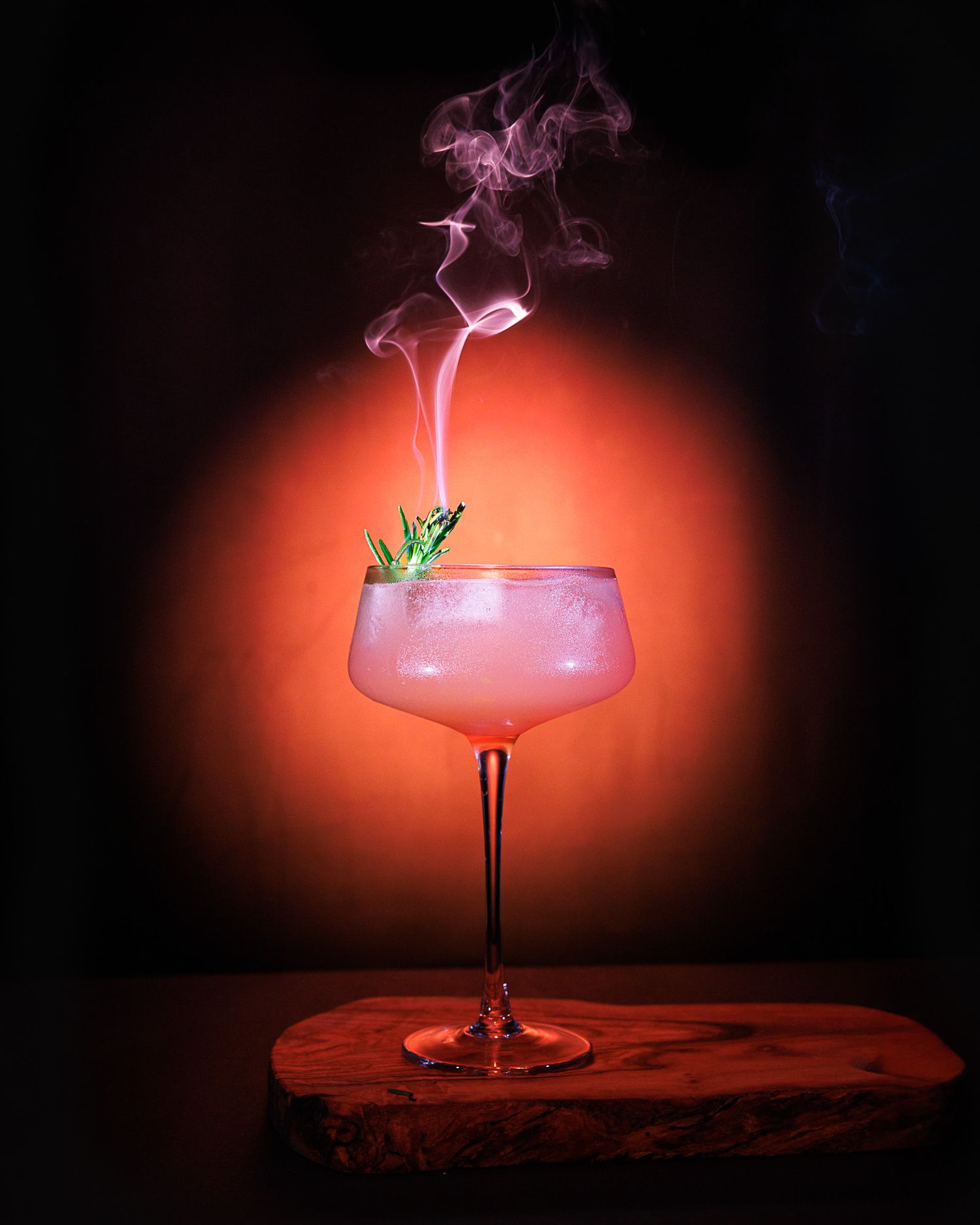 A smoky pink cocktail in a coupe glass garnished with fresh herbs on a wooden board against a red backdrop.